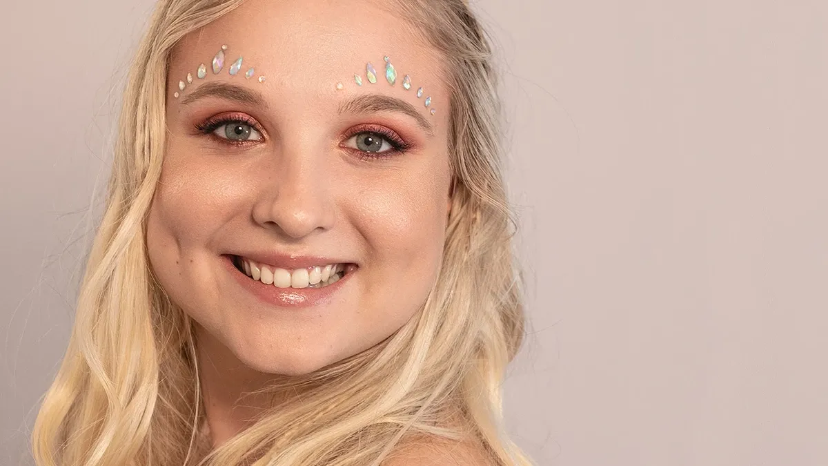 Smilende blond kvinne med kobberfarget øyenskygge og rhinestones på toppen av øyenbrynene.