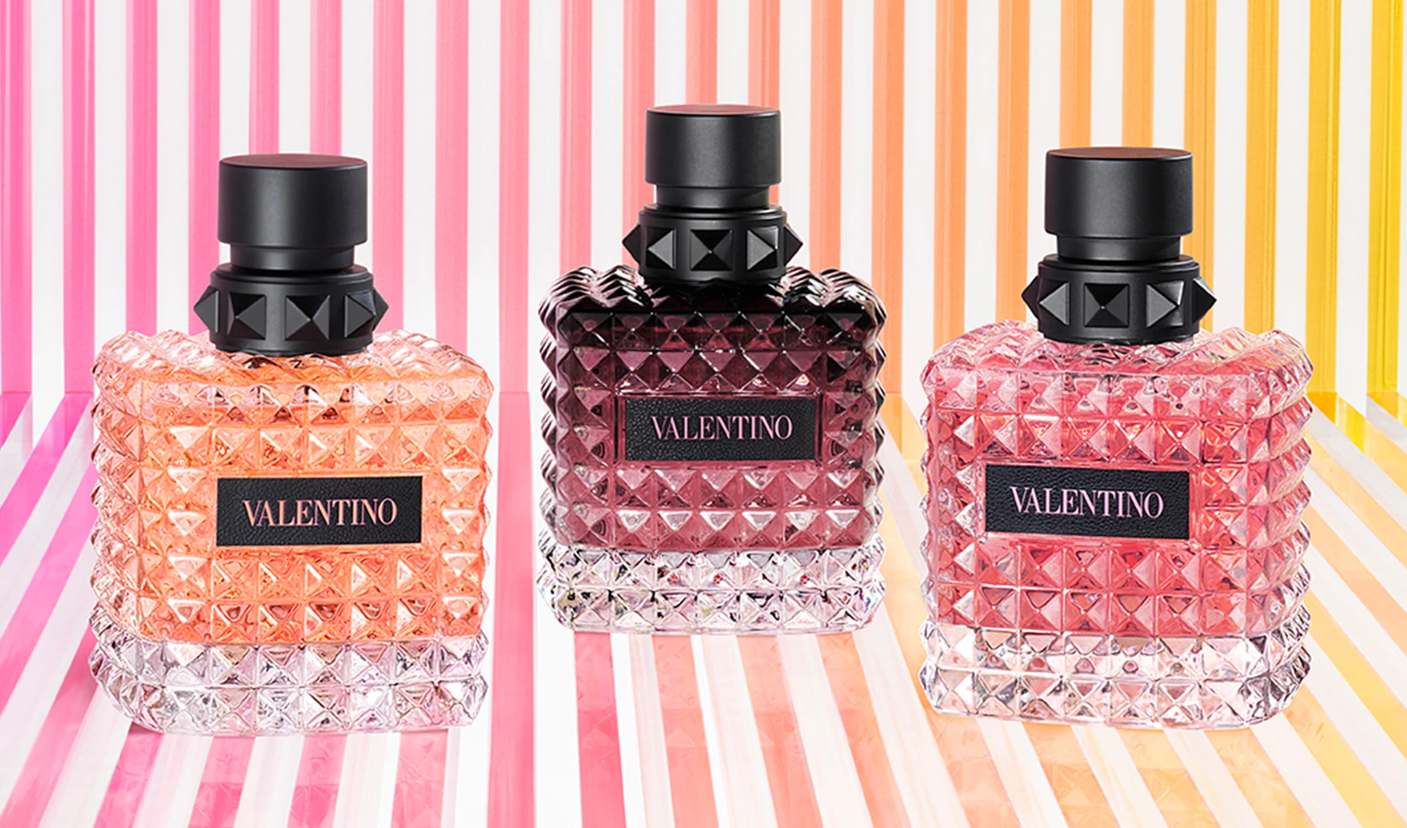 Tre forskjellige parfymeflasker fra Valentino på en stripete bakgrunn i rosa, oransje og gul.