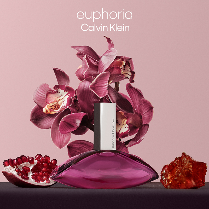 Calvin Klein-parfymflaska märkt "Euphoria". Flaskan är lila och bakom den finns purpurfärgade orkidéer. Framför finns ett granatäpple och en orange sten.