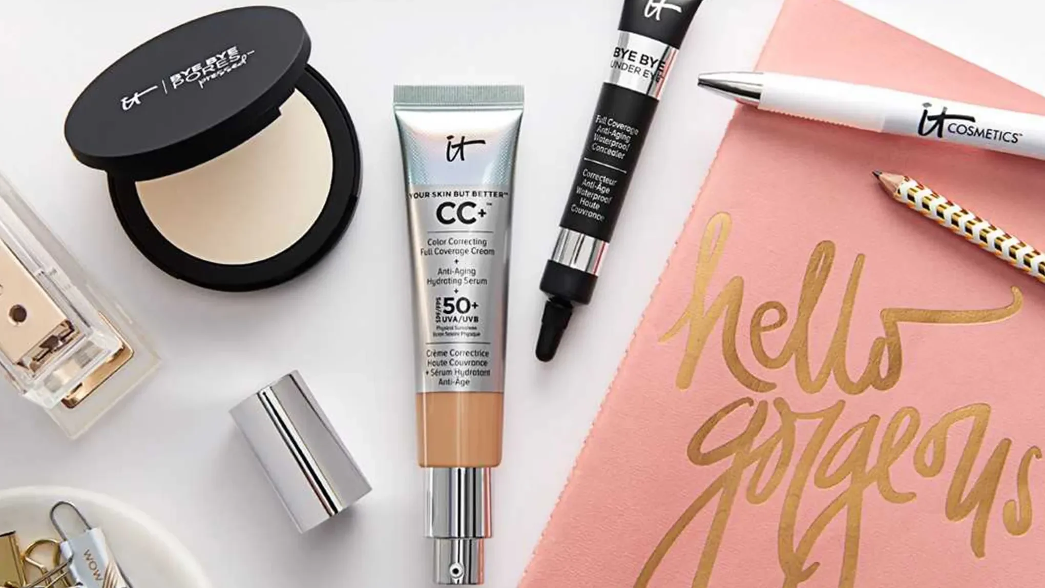 Flere produkter fra IT Cosmetics plassert på en hvit overflate, sammen med en rosa duk med teksten "hello gorgeous" i gull.