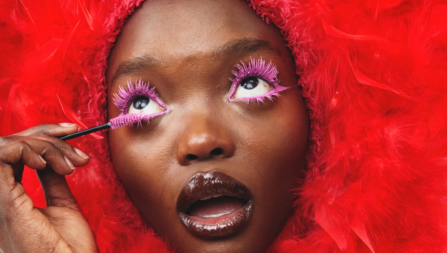 Nærbilde av en person som påfører rosa mascara på de nedre øyevippene. Øynene er innrammet med intens rosa øyesminke og mascara, leppene har blank lipgloss og ansiktet er omgitt av røde fjær.