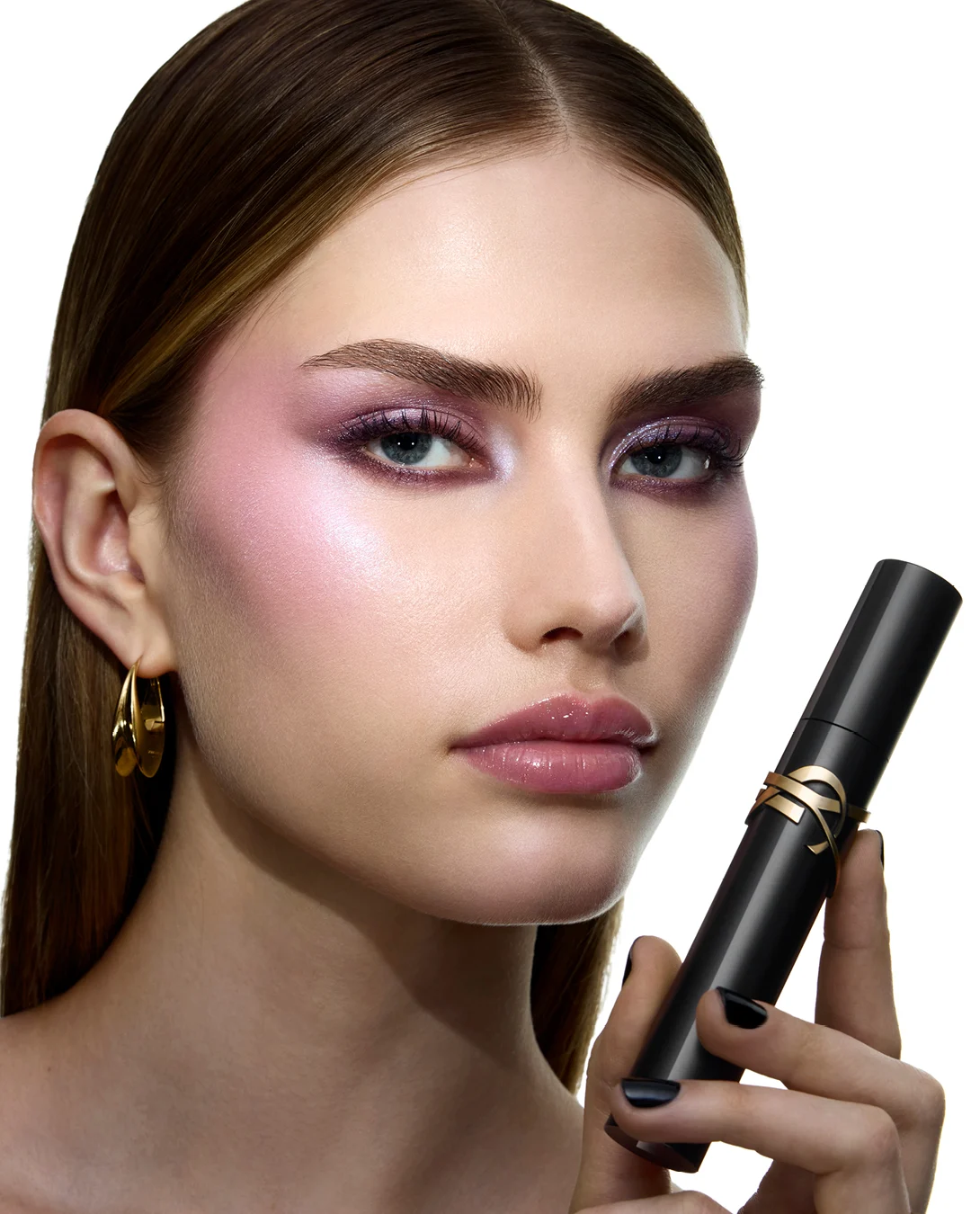 Närbild av en person med skimrande lila ögonmakeup, rosa highlighter och glansiga läppar. Personen håller en svart Yves Saint Laurent-produkt med guldfärgad YSL-logotyp och bär guldfärgade örhängen mot ljus bakgrund.