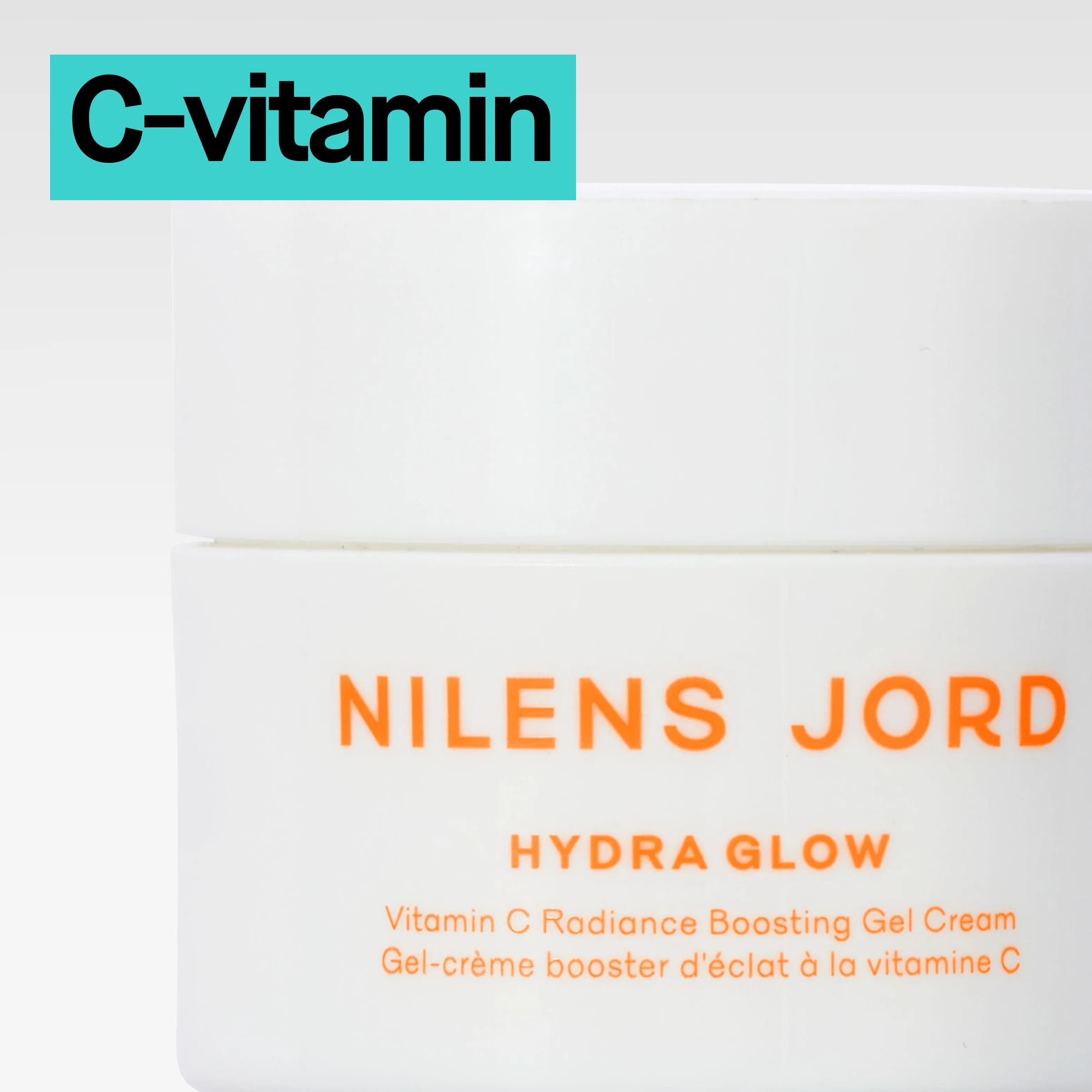 Hvit krukke med oransje tekst «Nilens Jord Hydra Glow Vitamin C Radiance Boosting Gel Cream», fotografert i nærbilde mot lys bakgrunn med turkis etikett merket «C-vitamin».