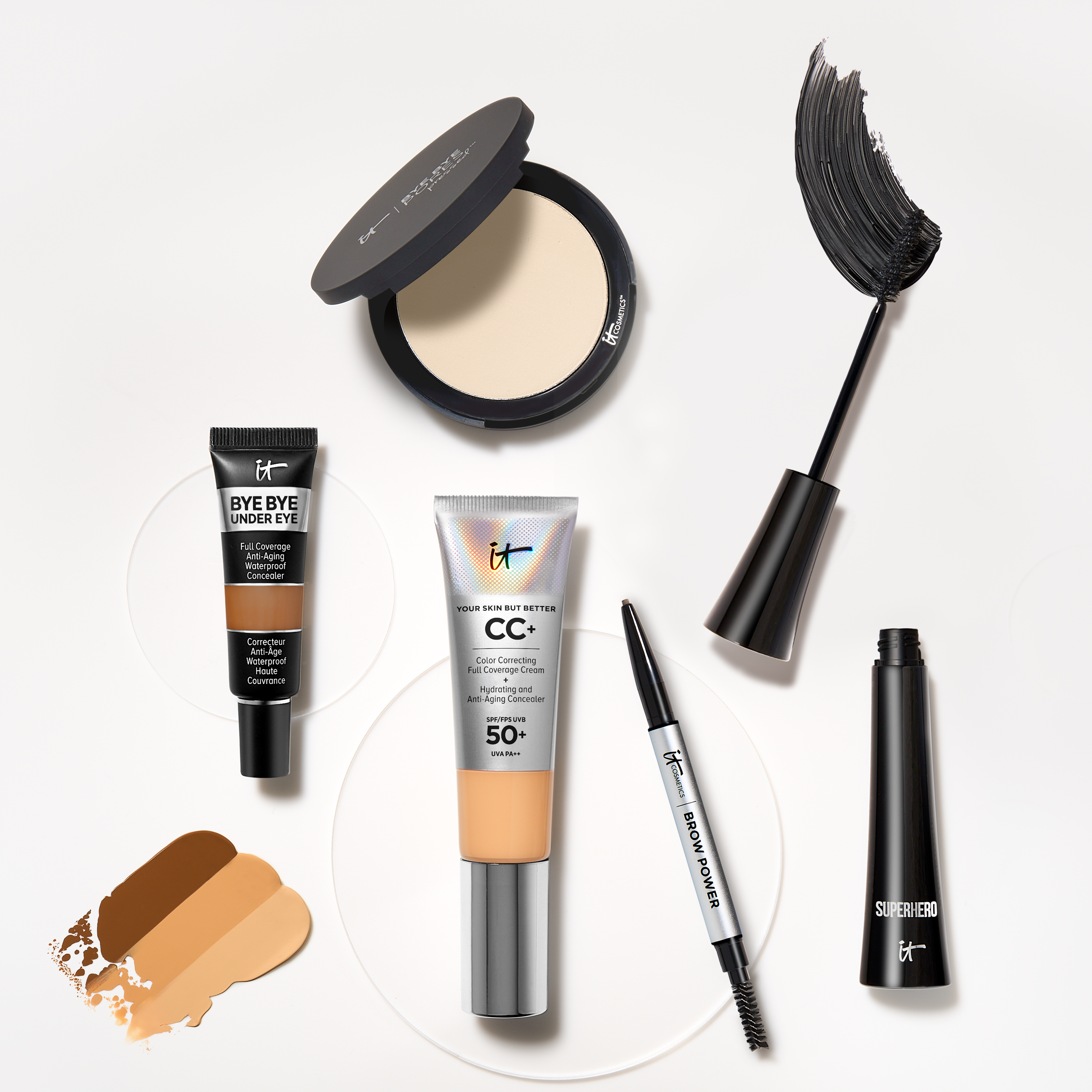 Useita IT Cosmeticsin meikkituotteita. Tuotteet on järjestetty vaalealle pinnalle, jossa on värinäytteitä peitevoiteesta ja CC+-voiteesta.