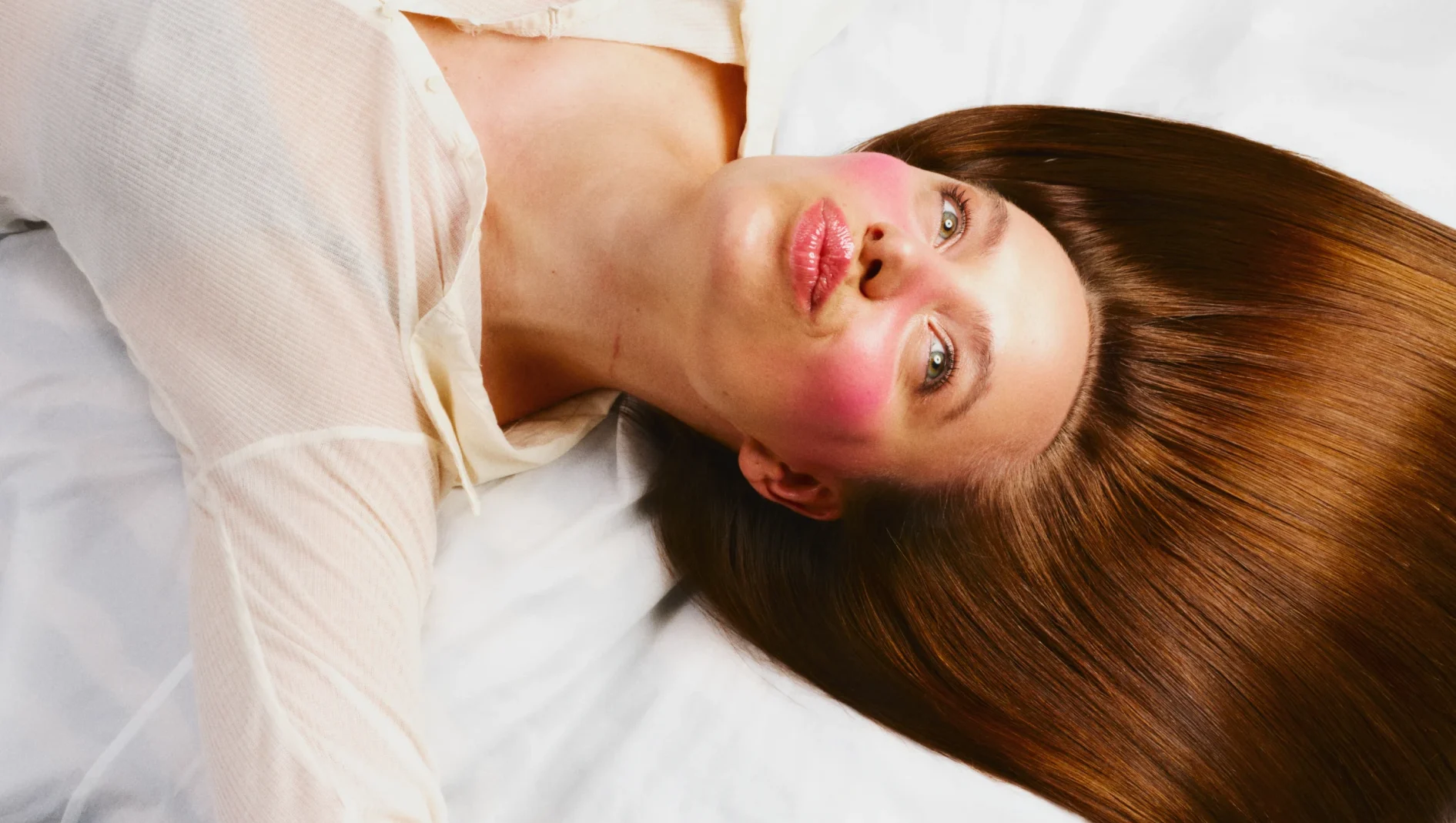 En person ligger på et hvitt underlag med langt, rett og glansfullt brunt hår spredt ut til siden. Ansiktet har rosa blush og blanke rosa lepper.