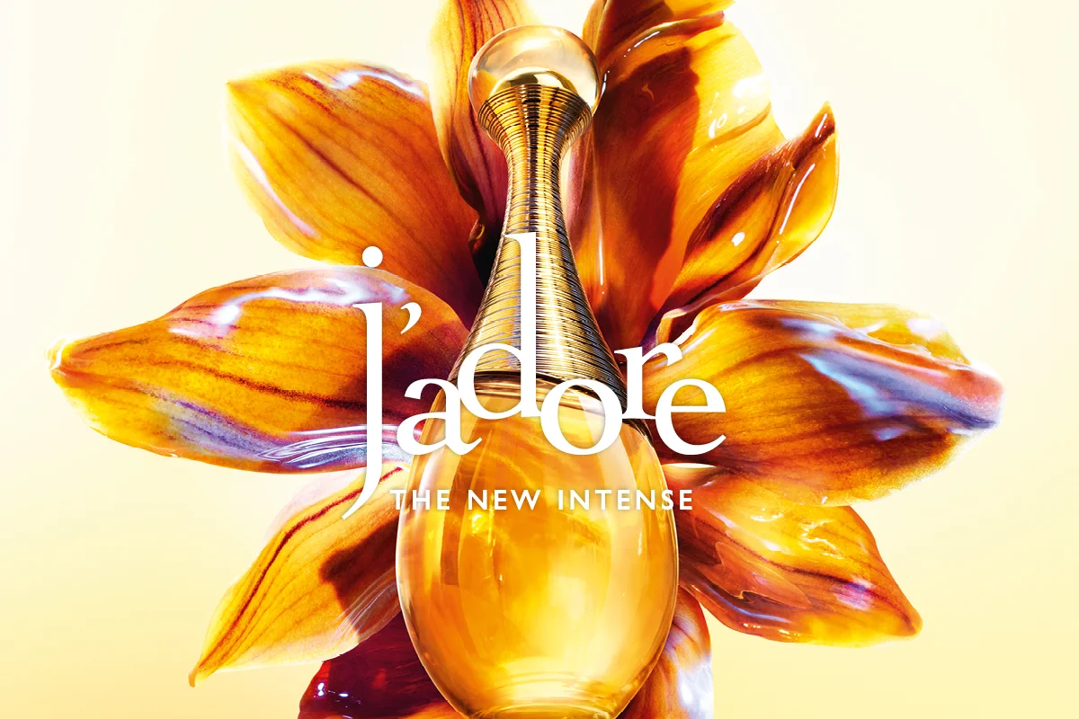 Dior J’adore parfymflaska i gyllene ton placerad framför en stor blomma i orange och gula nyanser med glansig yta. Text i bilden: "J’adore" och "THE NEW INTENSE".