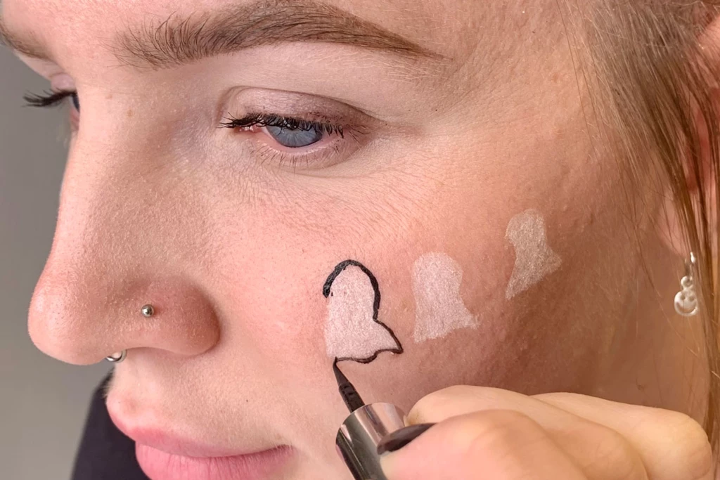 Person som tegner en hvit spøkelsesform på kinnet med svart flytende eyelinerpenn.