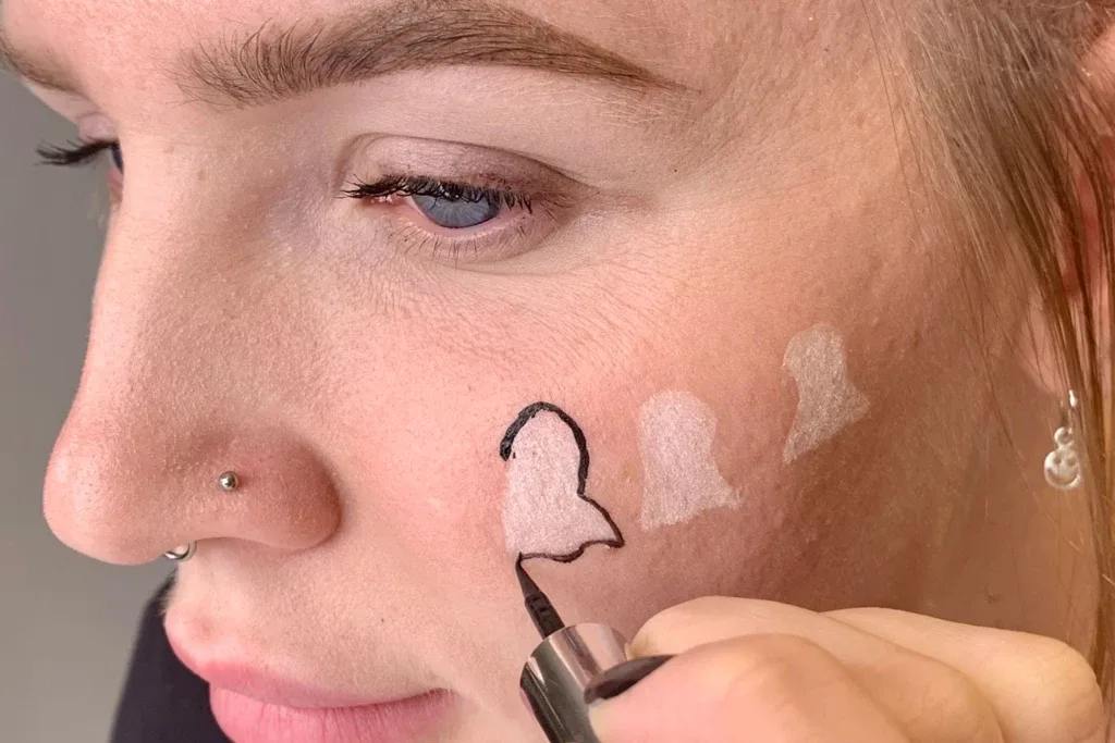 Henkilö rajaa poskelleen valkoisen haamumuodon nestemäisellä, mustalla eyeliner-kynällä.