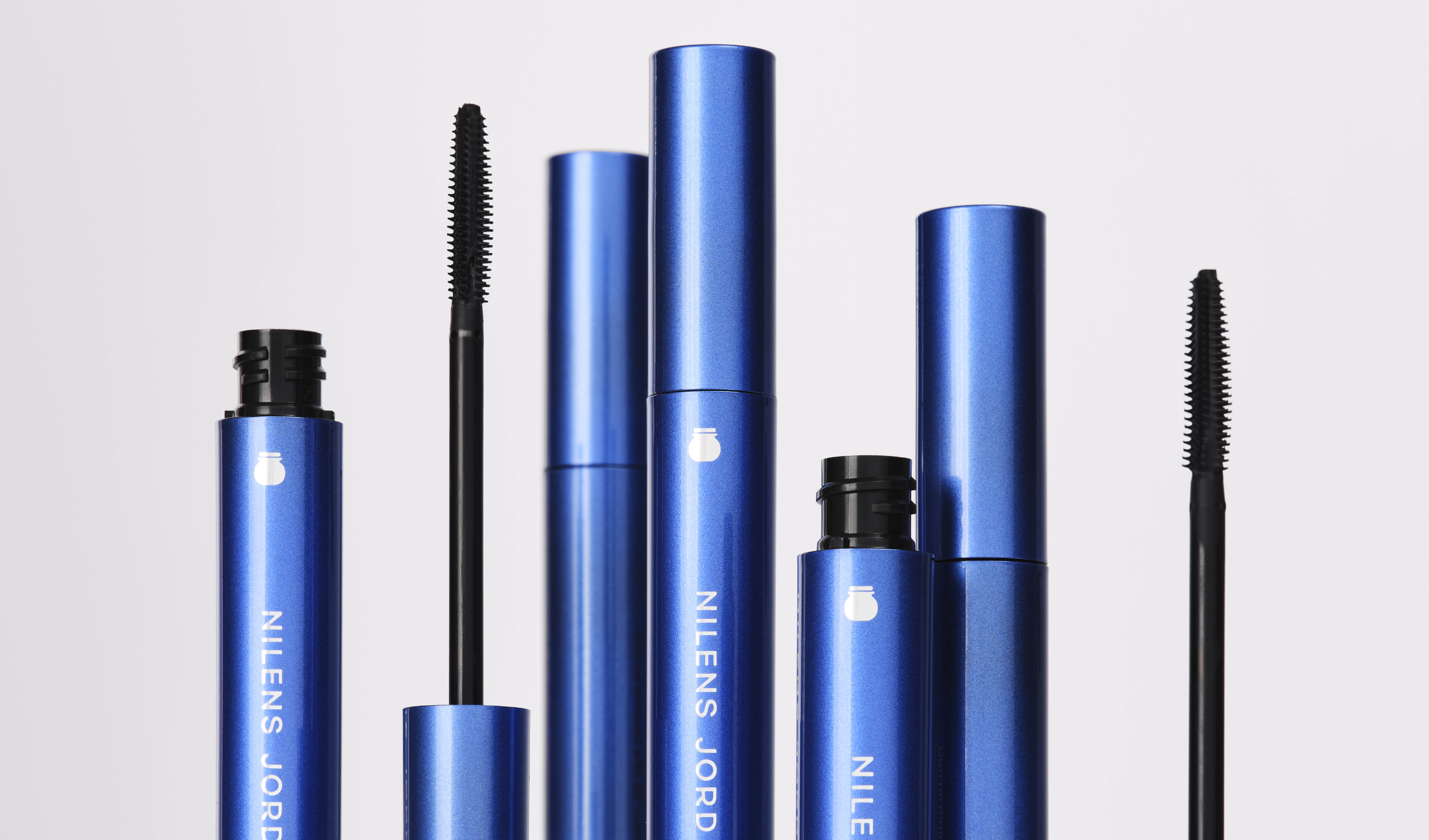 Blå mascaraer fra Nilens Jord vises åpne med synlige mascarabørster mot lys grå bakgrunn. Emballasjen har hvit tekst med varemerket NILENS JORD og viser mascara for volum og definisjon av vippene.