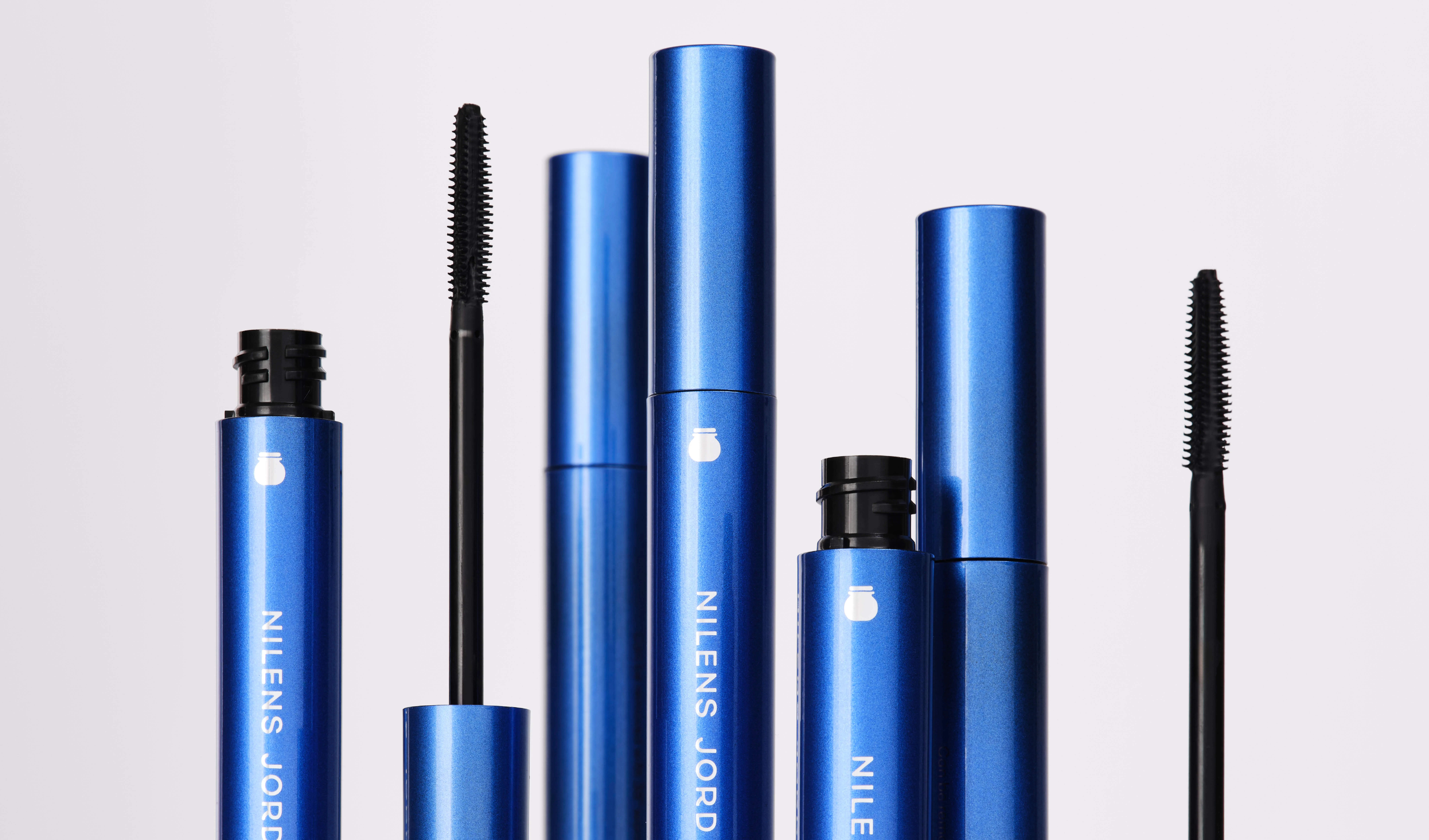 Blå mascaraer fra Nilens Jord vises åpne med synlige mascarabørster mot lys grå bakgrunn. Emballasjen har hvit tekst med varemerket NILENS JORD og viser mascara for volum og definisjon av vippene.