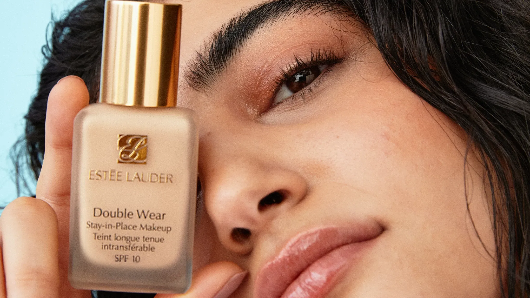 En kvinne med svart hår ser inn i kameraet og holder et produkt fra Estée Lauder foran øyet sitt.