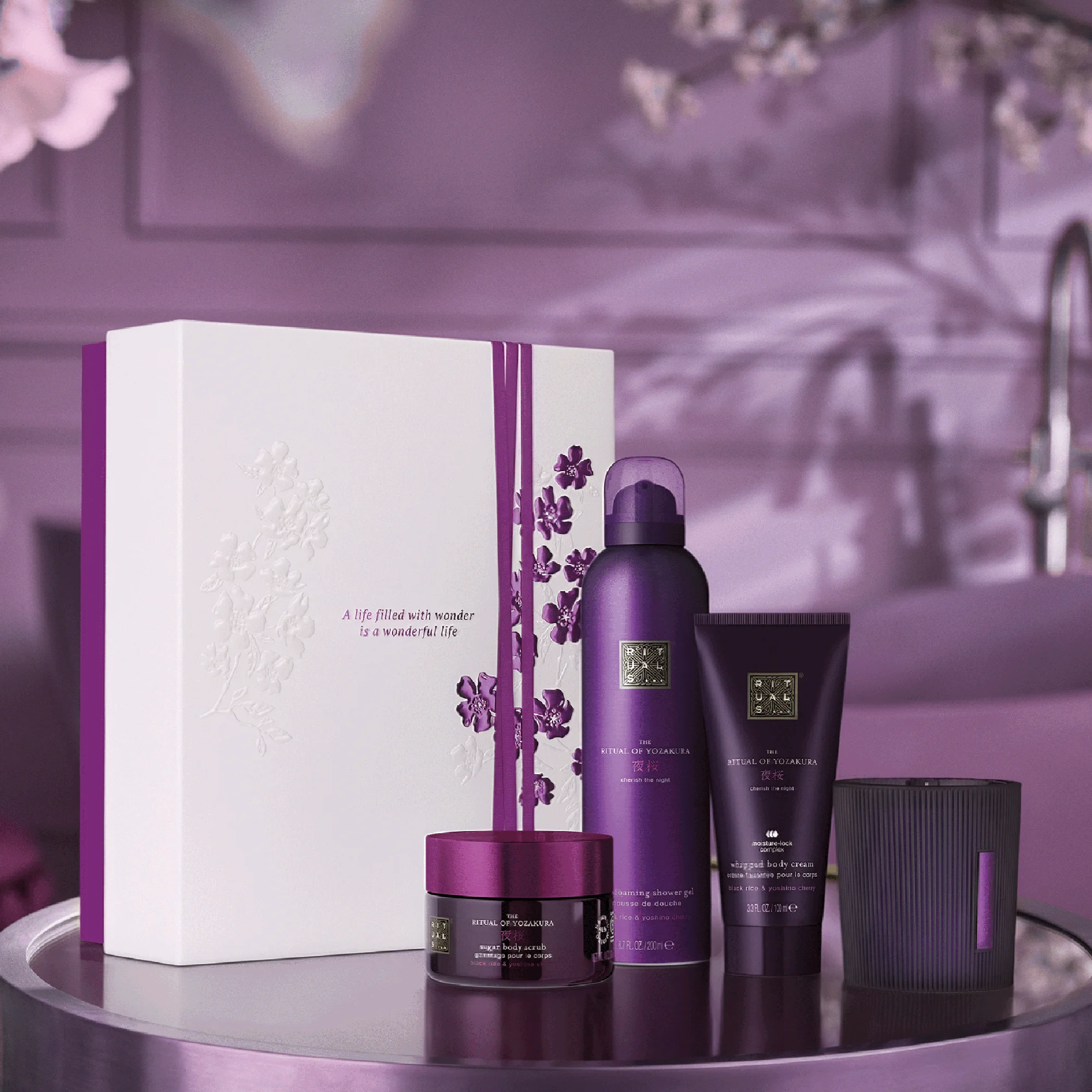 Presentset från Rituals The Ritual of Yozakura med foaming shower gel, whipped body cream, sugar body scrub och doftljus står bredvid en vit ask med blomdetaljer. På asken syns texten “A life filled with wonder is a wonderful life”.