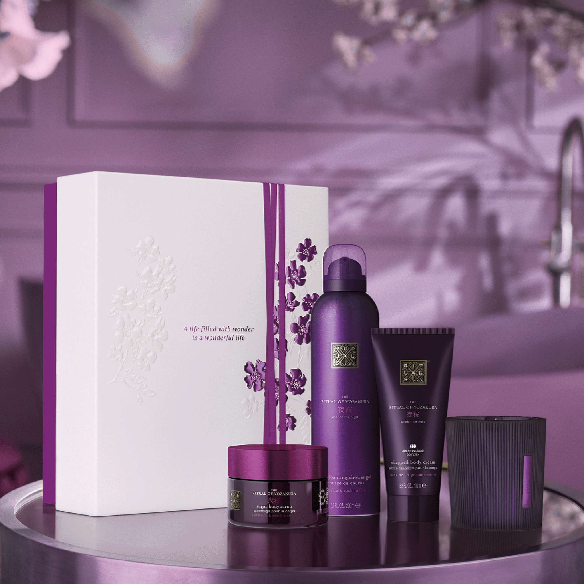 Gavesett fra Rituals The Ritual of Yozakura med foaming shower gel, whipped body cream, sugar body scrub og duftlys står ved siden av en hvit eske med blomsterdetaljer. På esken står teksten “A life filled with wonder is a wonderful life”.