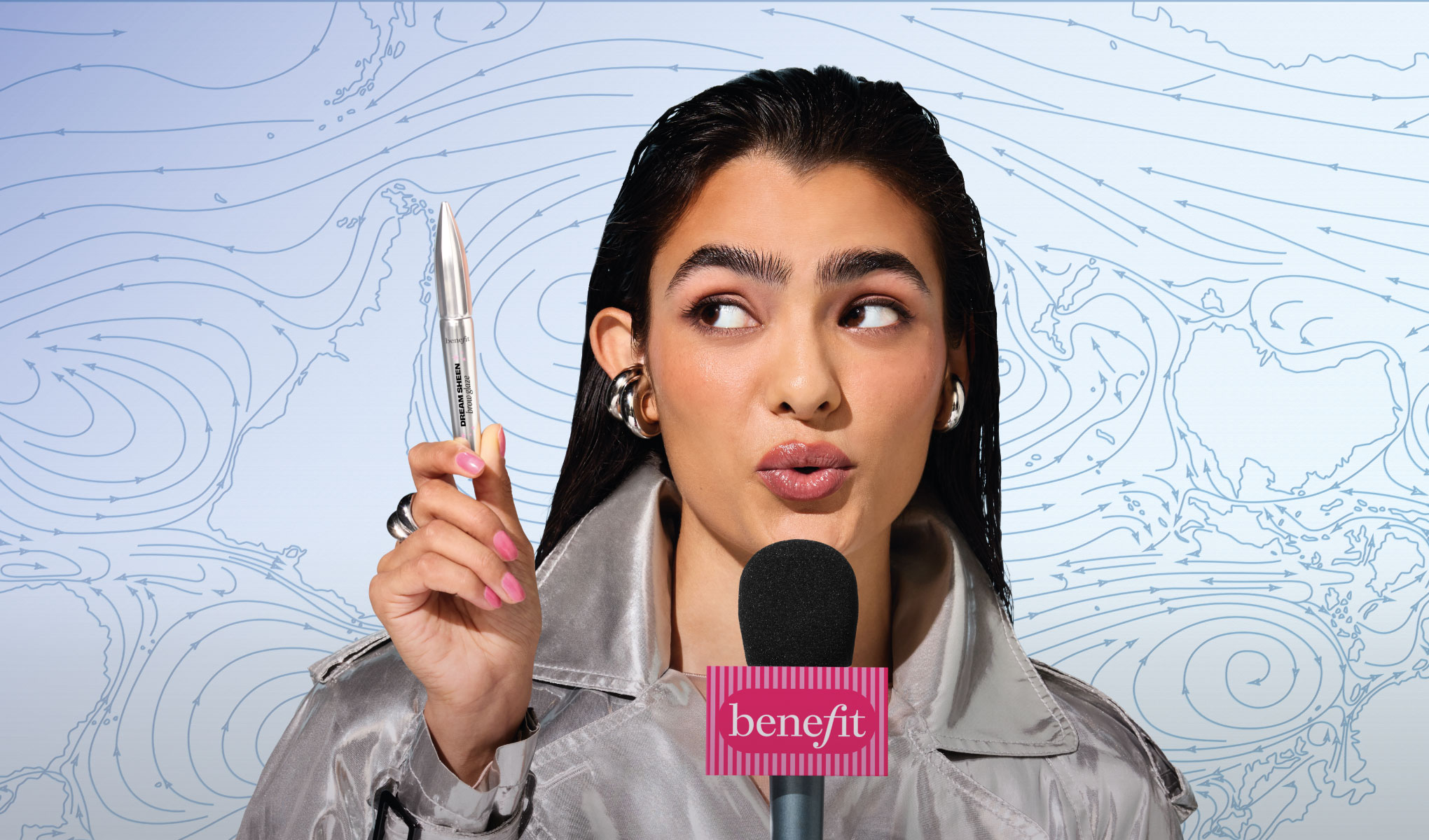 Person holder opp Benefit Dream Screen mascara og snakker i en mikrofon med teksten “benefit”, foran en bakgrunn med kartlignende linjemønster. Produktet er tydelig synlig sammen med sminke som mascara og glansfulle lepper.