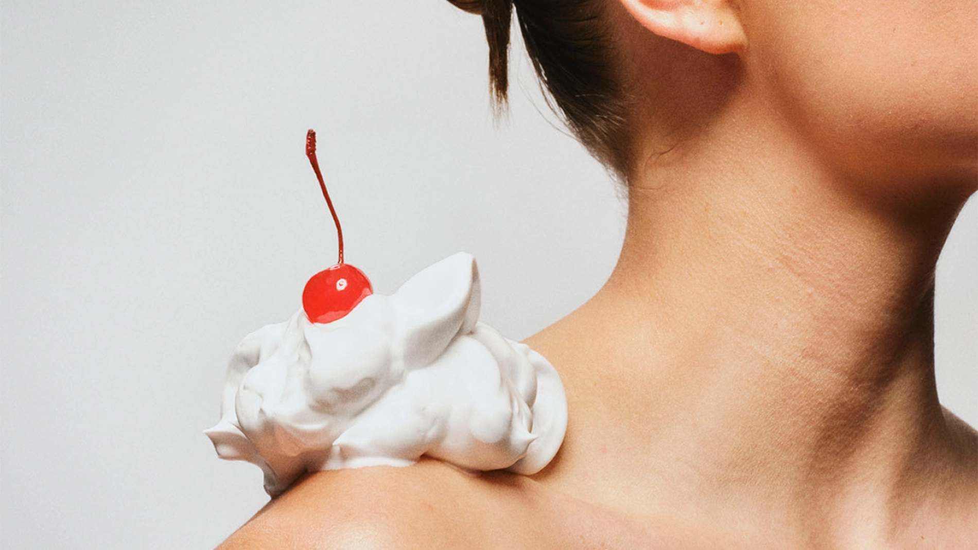 Nærbilde av en skulder med oppstrammende krem formet dekorativt og toppet med et kirsebær – en kreativ kampanjebilde som uttrykker fasthet, fylde og luksuriøs hudpleie.