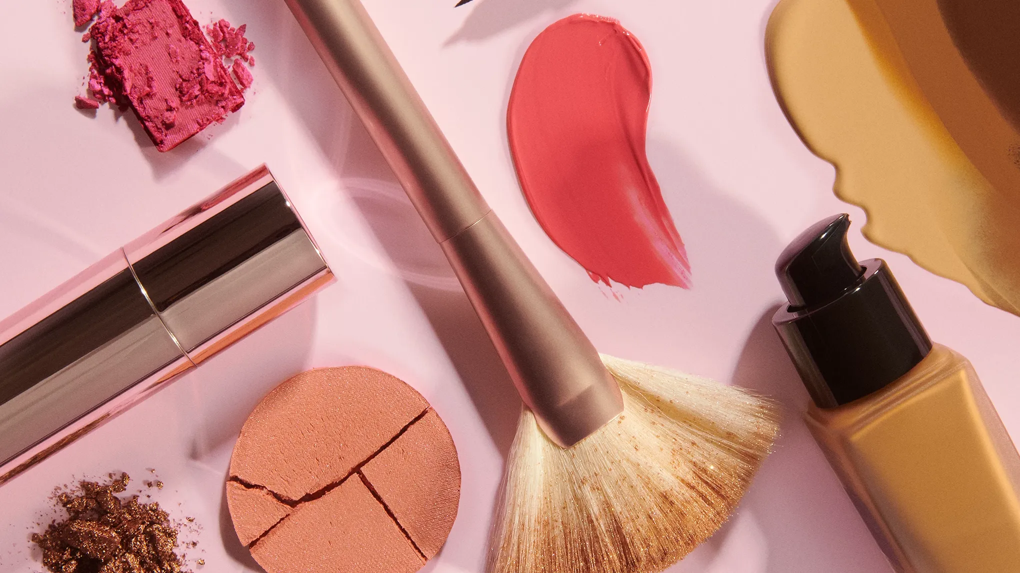 Sminkprodukter inklusive borste, läppstift, foundation och ögonskugga ligger på en rosa bakgrund.