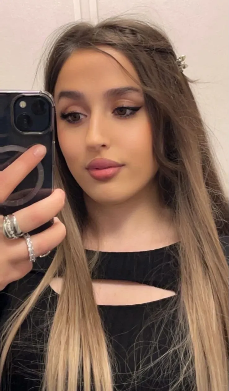 Porträtt av person som tar en spegelselfie med mobiltelefon. Personen har långt rakt hår med mittbena och hårspänne på sidan, markerad eyeliner och rosa läppar. Svart topp med utskärning vid halsringningen samt ringar på fingrarna syns mot en ljus inomhusbakgrund.