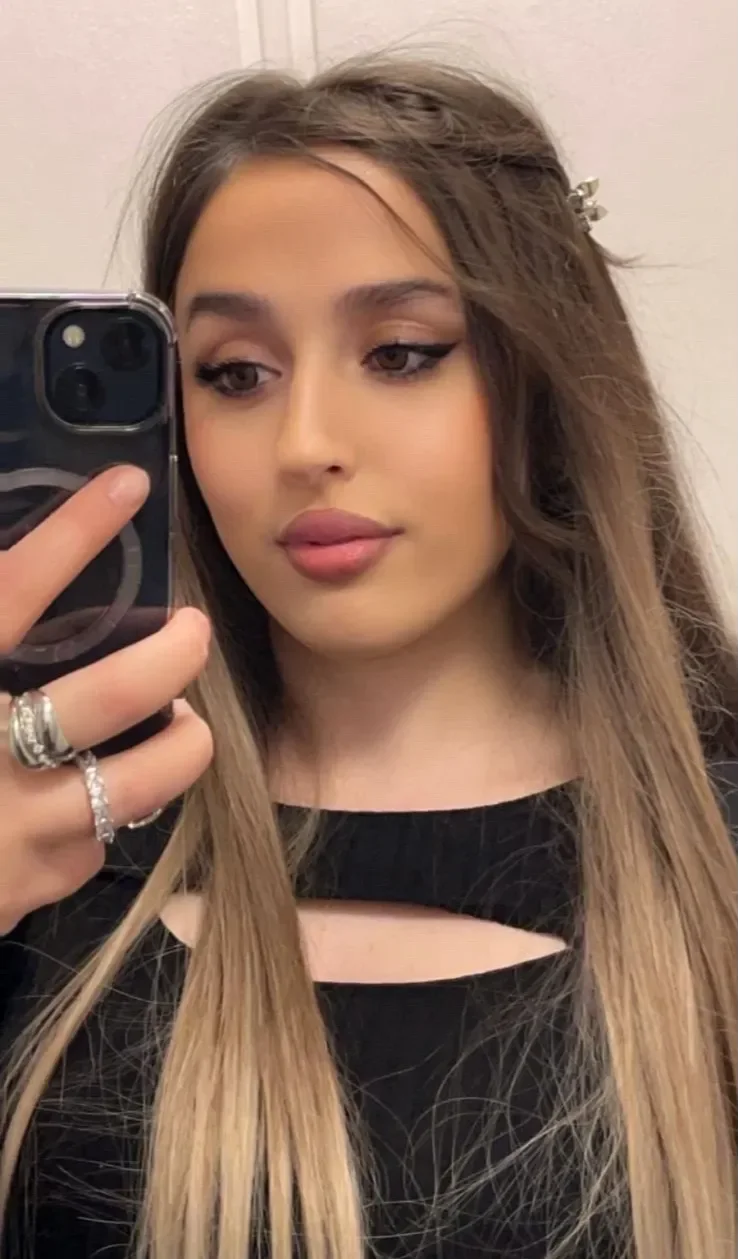 Porträtt av person som tar en spegelselfie med mobiltelefon. Personen har långt rakt hår med mittbena och hårspänne på sidan, markerad eyeliner och rosa läppar. Svart topp med utskärning vid halsringningen samt ringar på fingrarna syns mot en ljus inomhusbakgrund.