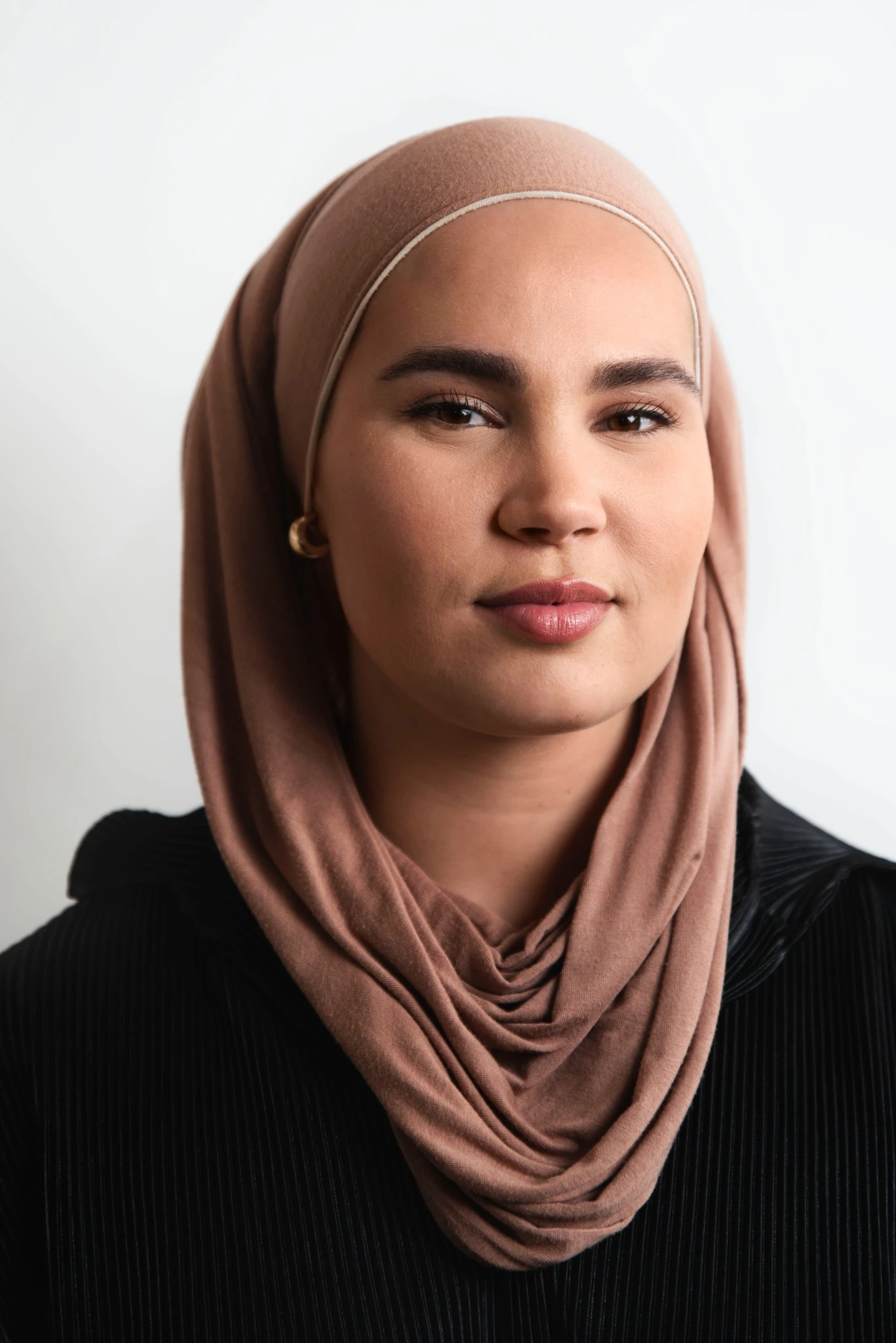 Portrett av person med brun hijab og svart plissert topp. Nærbilde mot lys bakgrunn med naturlig sminke, definerte øyenbryn og nøytrale lepper. Små øredobber er synlige.