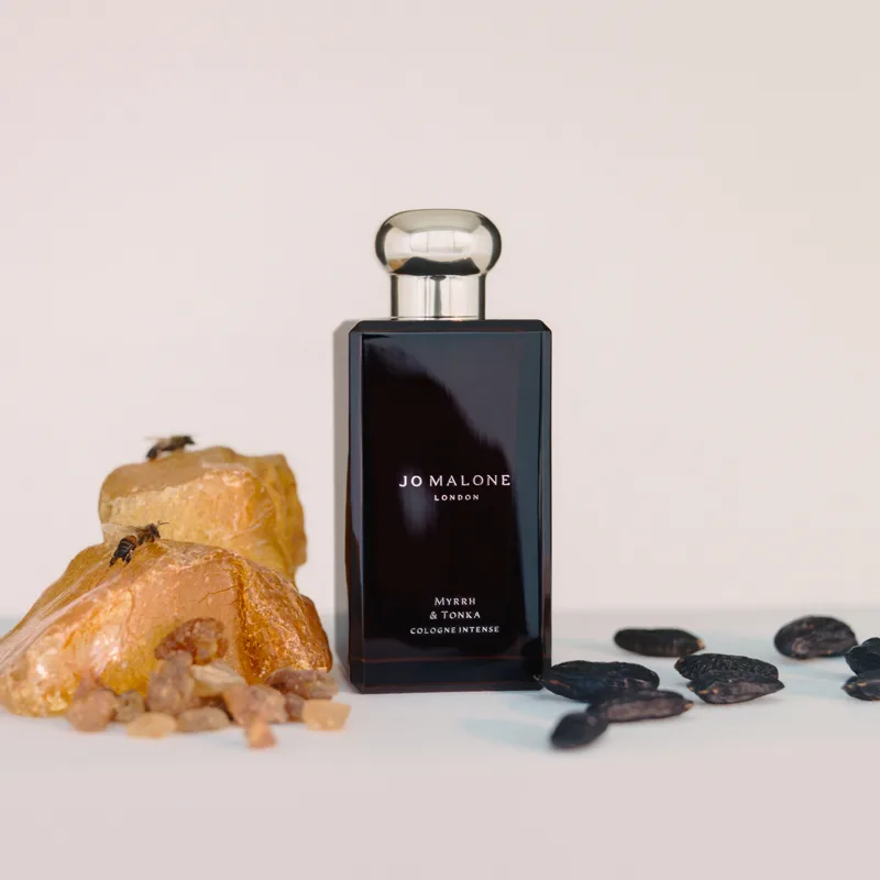 En svart parfymflaska från Jo Malone med texten "Myrrh & Tonka Cologne Intense" på en vit bakgrund. Bredvid flaskan finns myrrhstenar och tonkabönor.