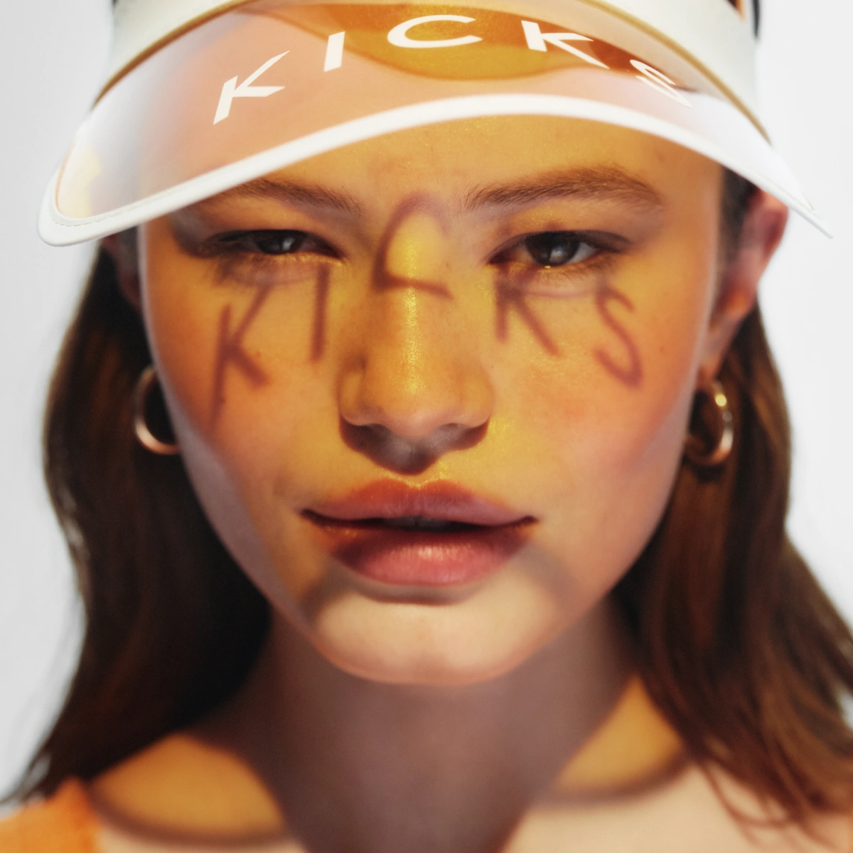 Henkilö, jolla on lippalakki tekstillä “KICKS” ja kasvoilla kirjaimia muodostava meikki.