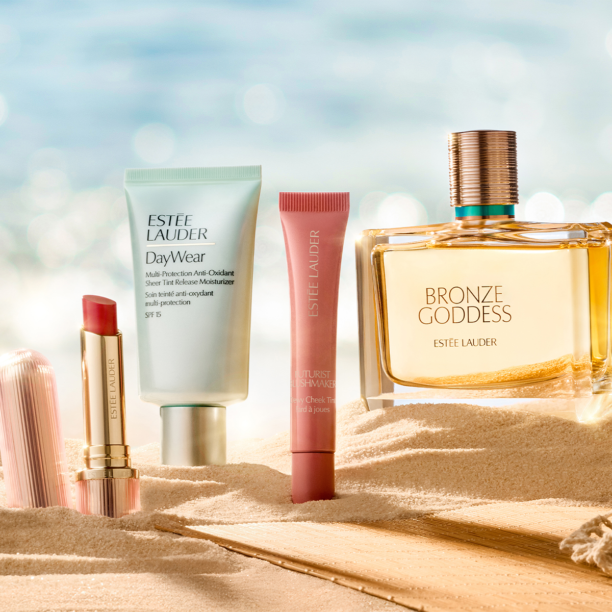 Fire Estée Lauder-produkter er plassert i sand foran en lys bakgrunn som minner om strand og hav. Bildet viser en leppestift fra Estée Lauder, DayWear Multi-Protection Anti-Oxidant Sheer Tint Release Moisturizer SPF 15, Futurist Soft Touch Brightening Skincealer samt parfymen Bronze Goddess med den synlige teksten «BRONZE GODDESS ESTÉE LAUDER».