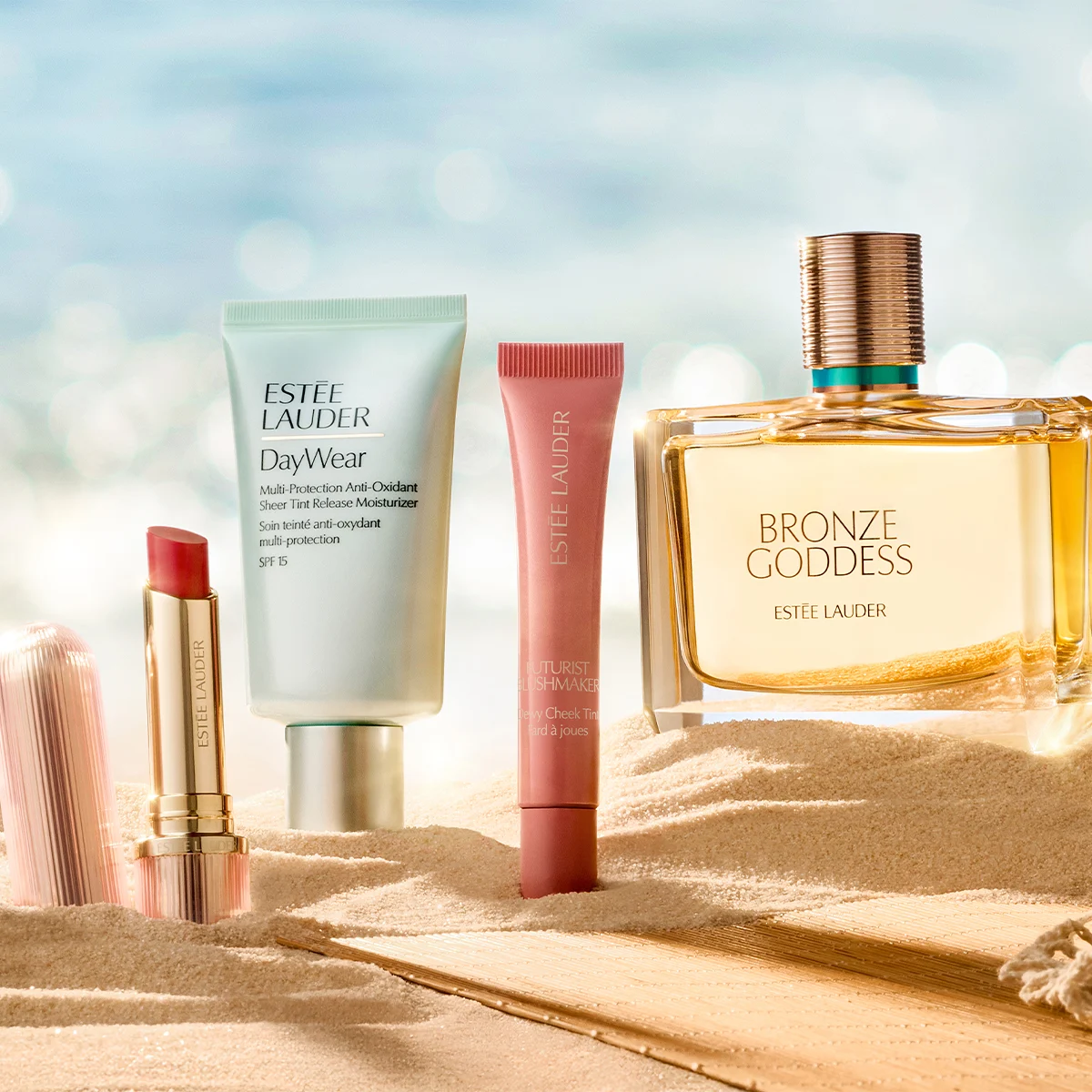 Neljä Estée Lauder -tuotetta on aseteltu hiekkaan vaalean ranta- ja merimaisemaa muistuttavan taustan eteen. Kuvassa näkyvät Estée Lauder -huulipuna, DayWear Multi-Protection Anti-Oxidant Sheer Tint Release Moisturizer SPF 15, Futurist Soft Touch Brightening Skincealer sekä Bronze Goddess -hajuvesi, jossa on näkyvä teksti ”BRONZE GODDESS ESTÉE LAUDER”.