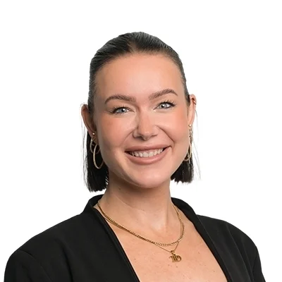 Porträtt av en leende person med mörk uppsatt frisyr, svart kavaj och guldsmycken mot ljusgrå bakgrund. Personen bär naturlig makeup med definierade bryn och nude läppfärg.