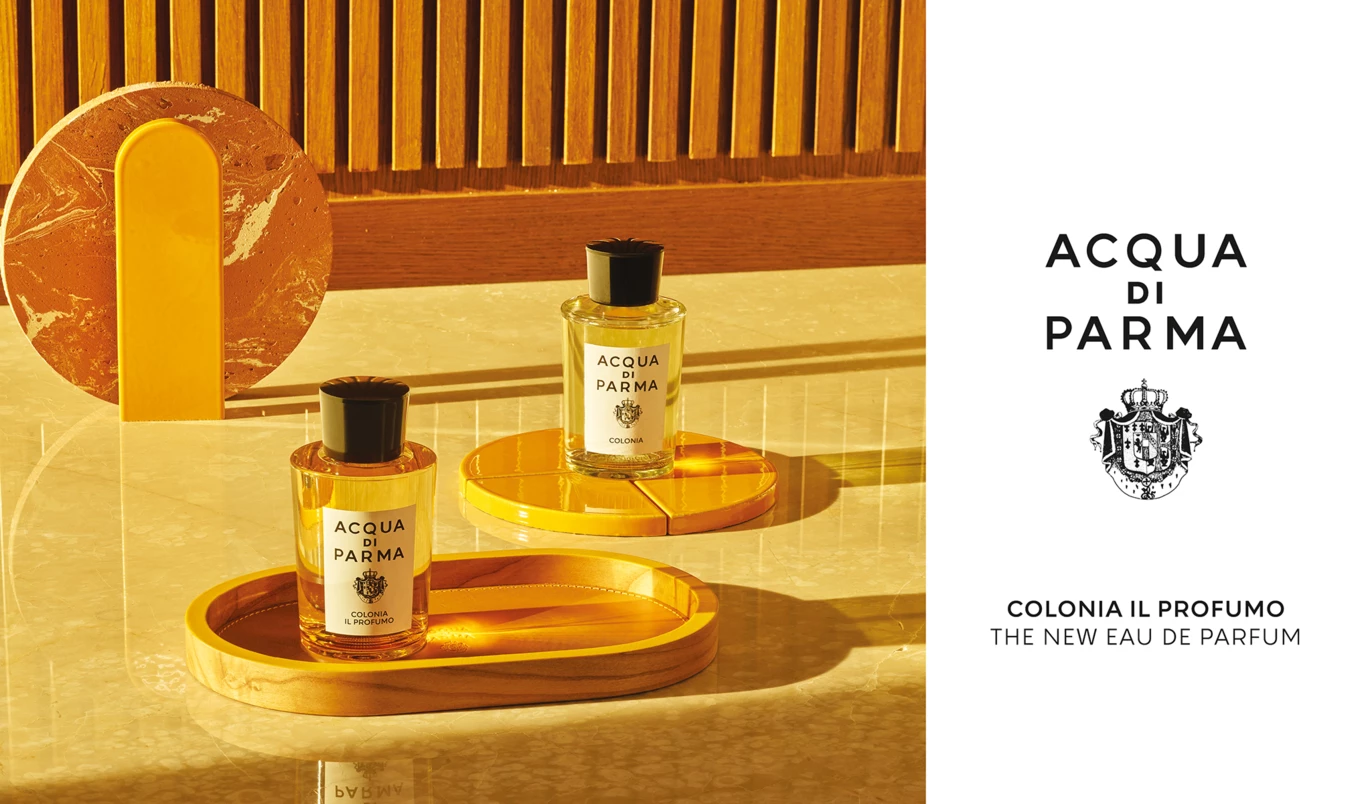 Acqua di Parma -hajuvesipullot keltaisilla tarjottimilla. Taustalla on marmorilaatta ja puupaneeli. Kuvan teksti on: ”Acqua di Parma Colonia Il Profumo The New Eau de Parfum”.