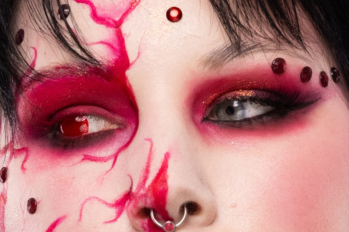 Porträtt med kreativ halloweenmakeup där halva ansiktet är täckt av rosa och röda målade ådror och ett skelettinspirerat mönster, kompletterat med röda dekorstenar och djup ögonmakeup.