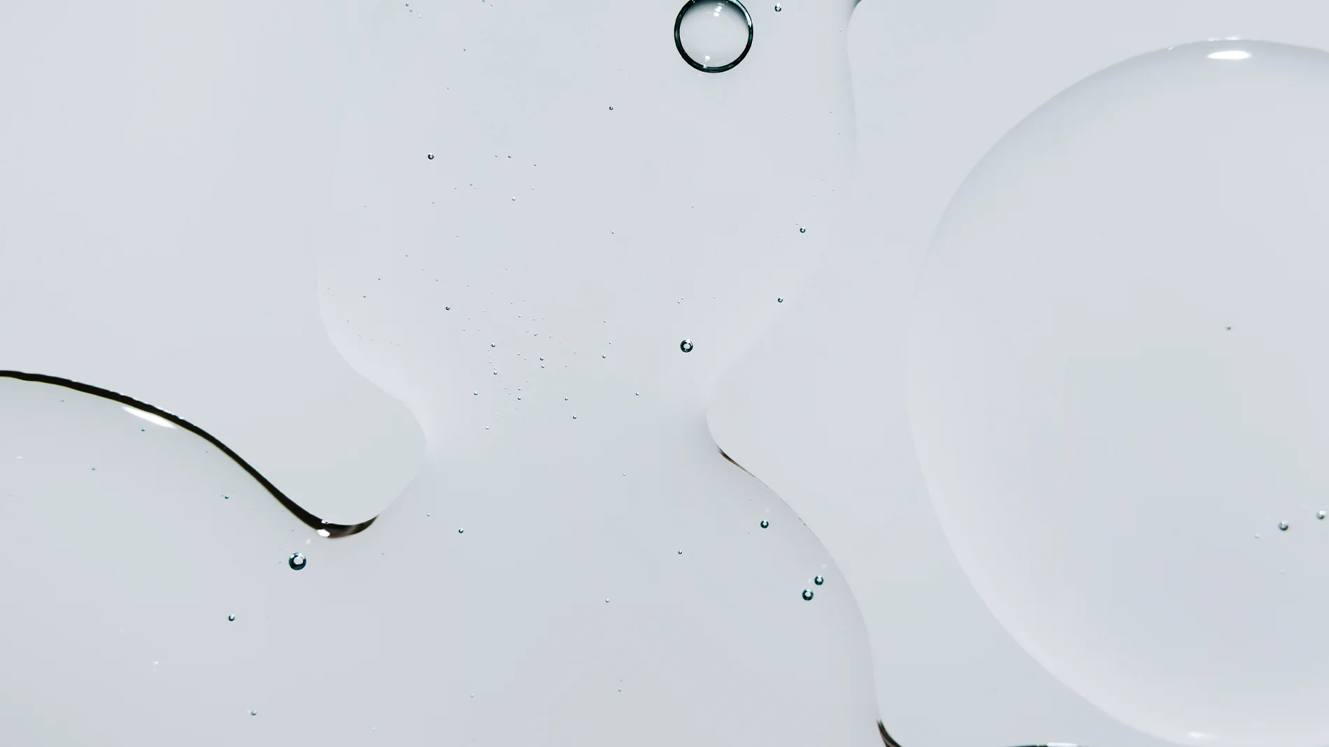 Lähikuva kirkkaasta ihonhoitotekstuurista, joka leviää pehmeisiin muotoihin vaalealla taustalla, pienillä pisaroilla ja ohuilla seerumireunoilla.