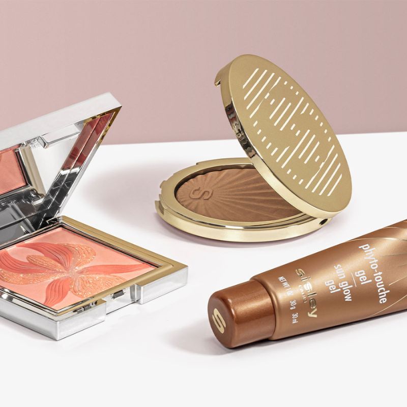 Tre sminkprodukter från Sisley placerade på en vit yta med en rosa bakgrund. En öppen palett med persikofärgad rouge, en öppen kompakt med bronzer samt en tub. Produkterna har förpackningar i guld och silver.