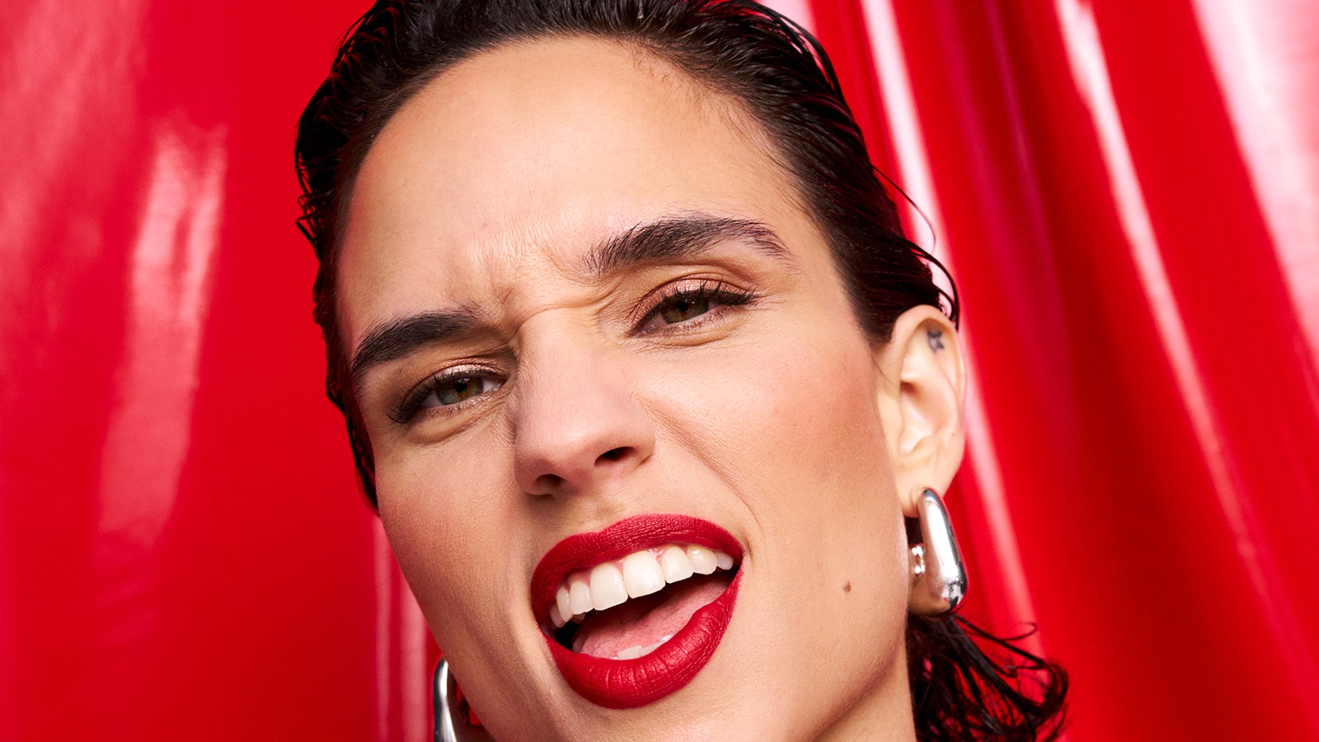 Nærbilde av en person med røde lepper, bakoverkjemmet hår og store sølvøredobber mot en blank rød bakgrunn. Et sterkt og selvsikkert uttrykk som gir en festlig og glamorøs følelse.