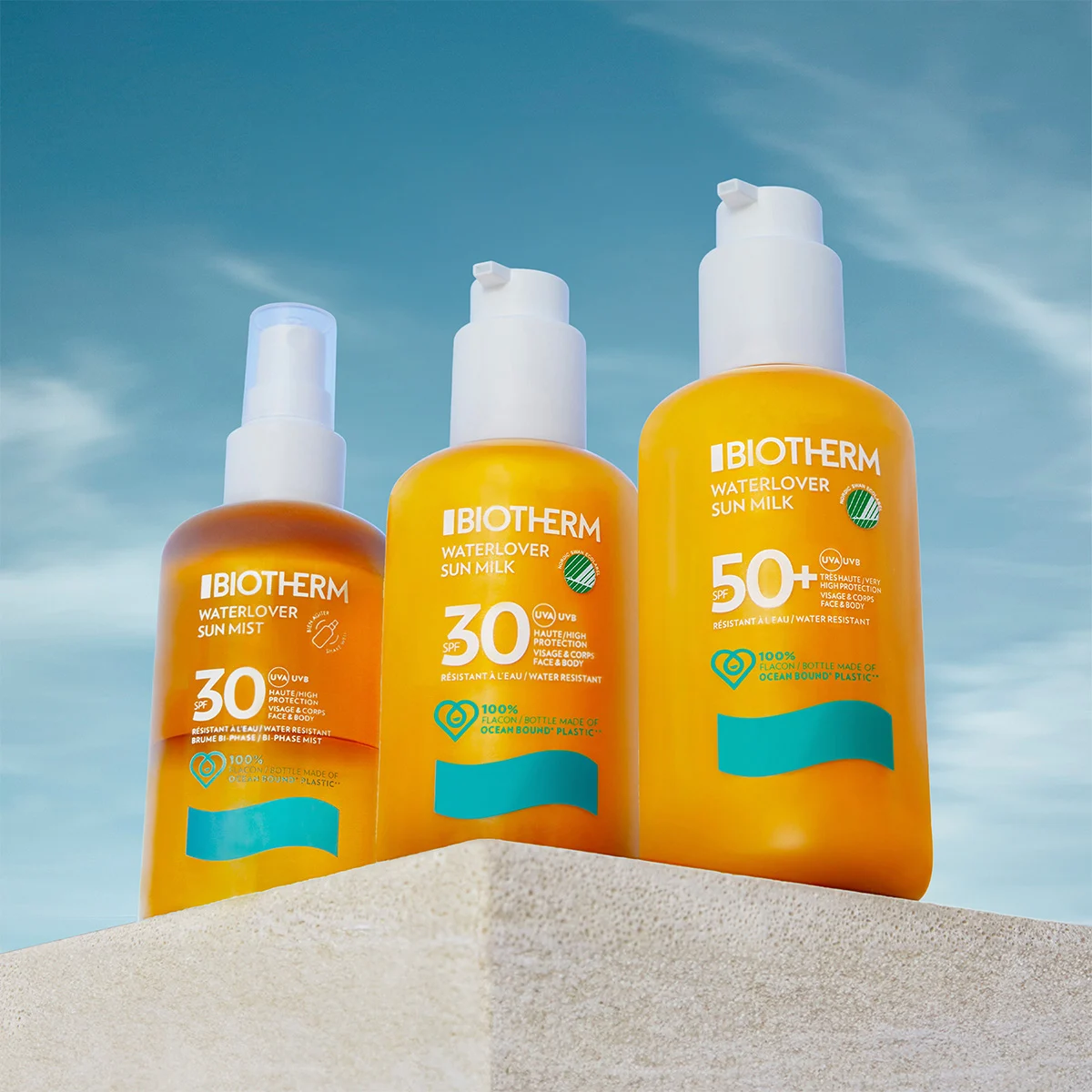 Tre solbeskyttelsesprodukter fra Biotherm er plassert på en beige plattform mot en blå himmel. Produktene inkluderer: en brun sprayflaske, en gul pumpeflaske og en større gul pumpeflaske.