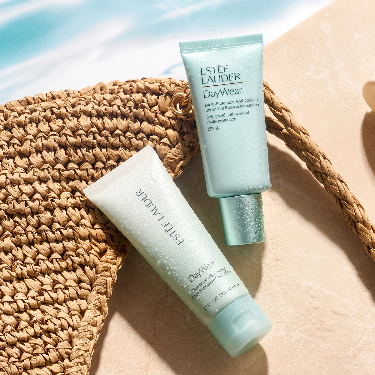 Kaksi Estée Lauder DayWear -ihonhoitotuotetta on aseteltu hiekanväriselle pinnalle punotun rantalaukun viereen. Tuubeissa näkyy vesipisaroita ja tekstit ”ESTÉE LAUDER DayWear Multi-Protection Anti-Oxidant Sheer Tint Release Moisturizer SPF 15” sekä ”ESTÉE LAUDER DayWear Glow Boosting Jelly Cleanser 150 ml”.