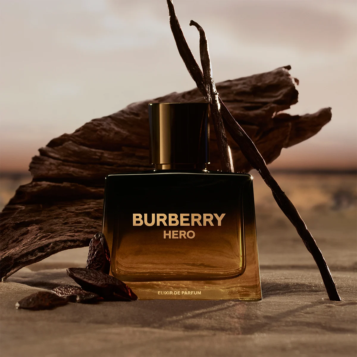 Burberry Hero Elixir de Parfum-flaske plassert på sand i solnedgang, omgitt av drivved og vaniljestenger.