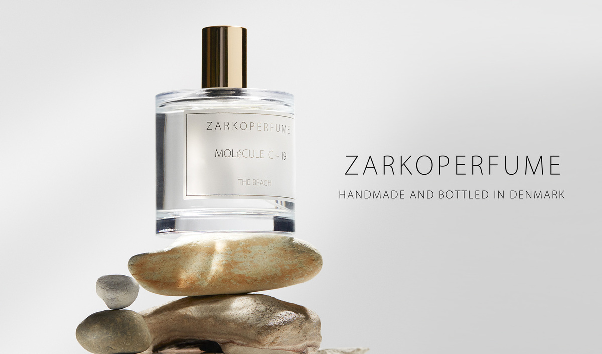 Hajuvesipullo Zarkoperfume. Pullo on läpinäkyvä ja siinä on kullanvärinen korkki, ja se on tasapainotettu kivipinon päällä. Pullon oikealla puolella on teksti ”ZARKOPERFUME Handmade And Bottled In Denmark” mustalla värillä vaaleanharmaalla pohjalla.