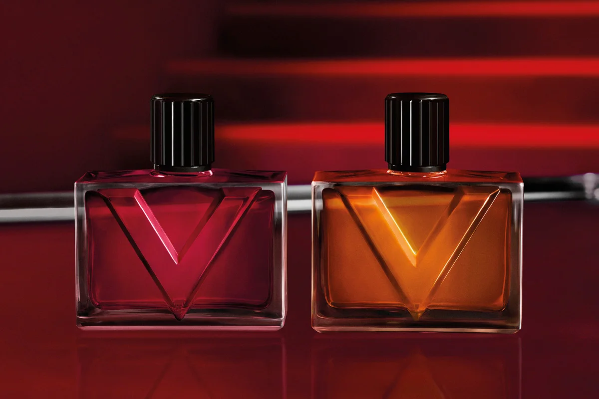 To rektangulære parfymeflasker i glass med svarte korker og en stor V-formet detalj på forsiden, én i dyp rødrosa og én i ravfarget oransje. Flaskene står mot en mørkerød bakgrunn med røde lysreflekser.