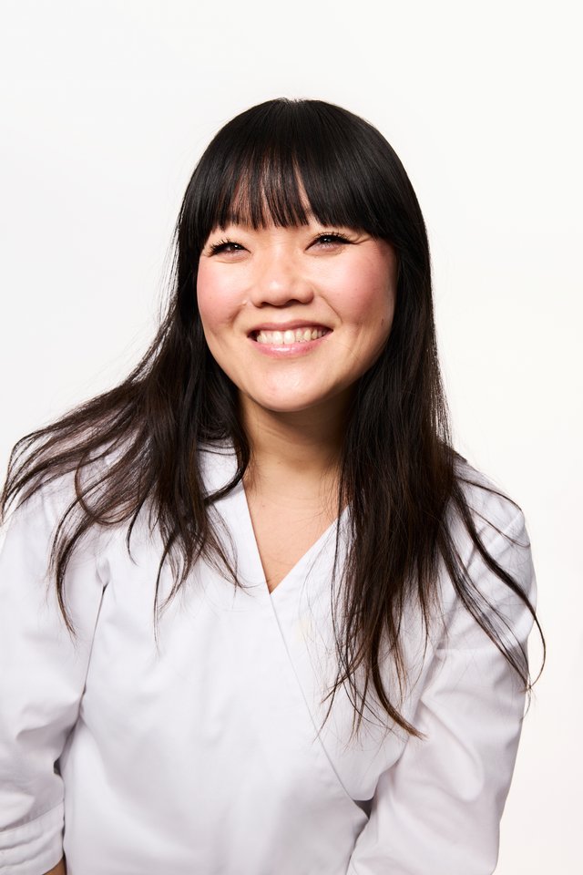 Mørkhåret, smilende person med pannelugg. Kledd i hvit frakk og bakgrunnen er hvit.