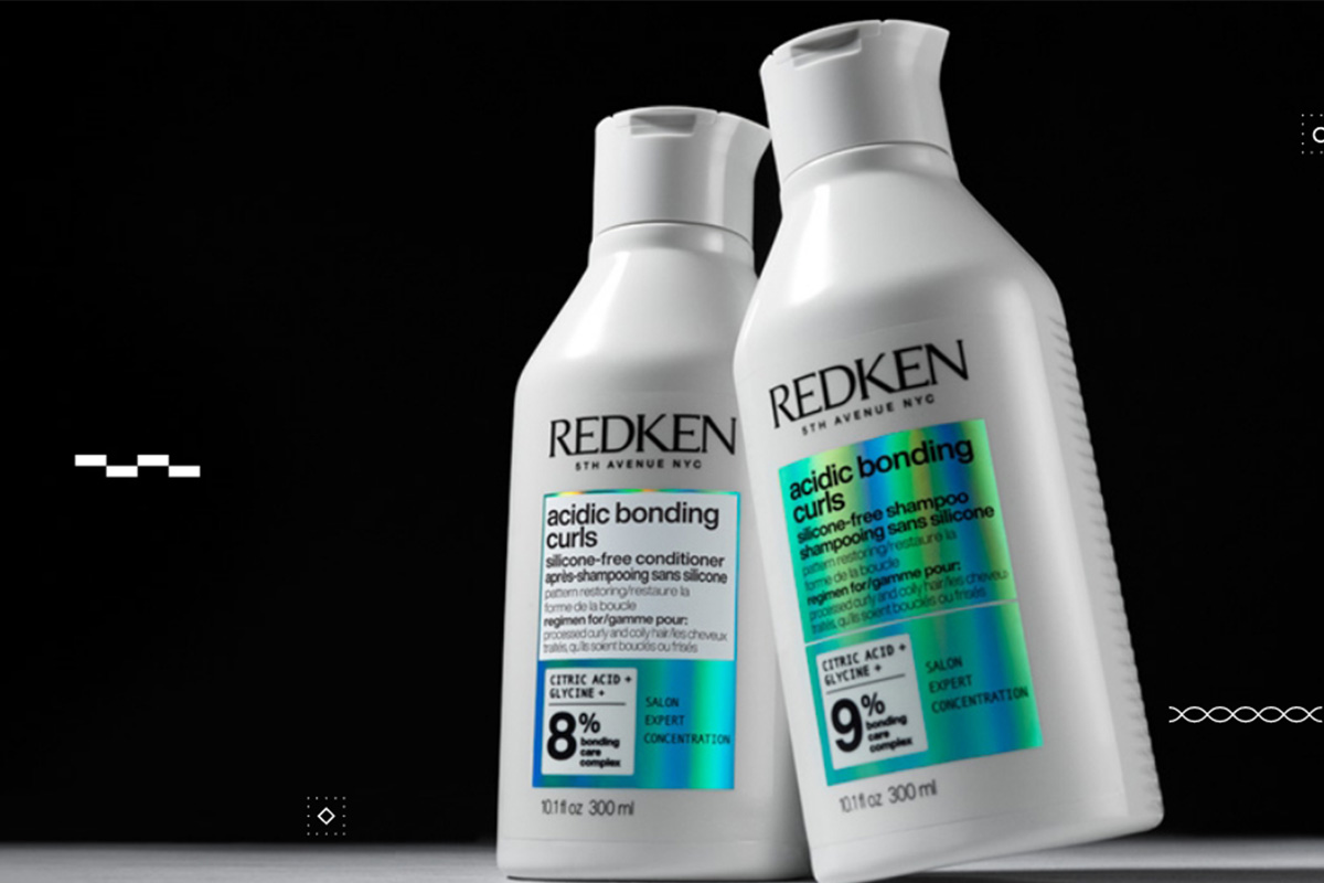 Kaksi valkoista Redken Acidic Bonding Curls -pulloa seisoo tummaa taustaa vasten. Toisessa pullossa on sininen ja vihreä etiketti ja toisessa sininen ja vihreä etiketti. Pullot on sijoitettu niin, että ne ovat toisiaan kohti.