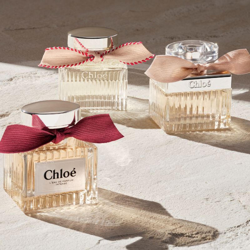 Tre parfymflaskor från Chloé. Ena flaskan är dekorerad med en mörkröd rosett och har en silverfärgad kork. De andra två är dekorerade med ljusrosa rosett. De är placerad på en ljus yta med mjuk belysning som skapar reflektioner. Bakgrunden är ljus och neutral.