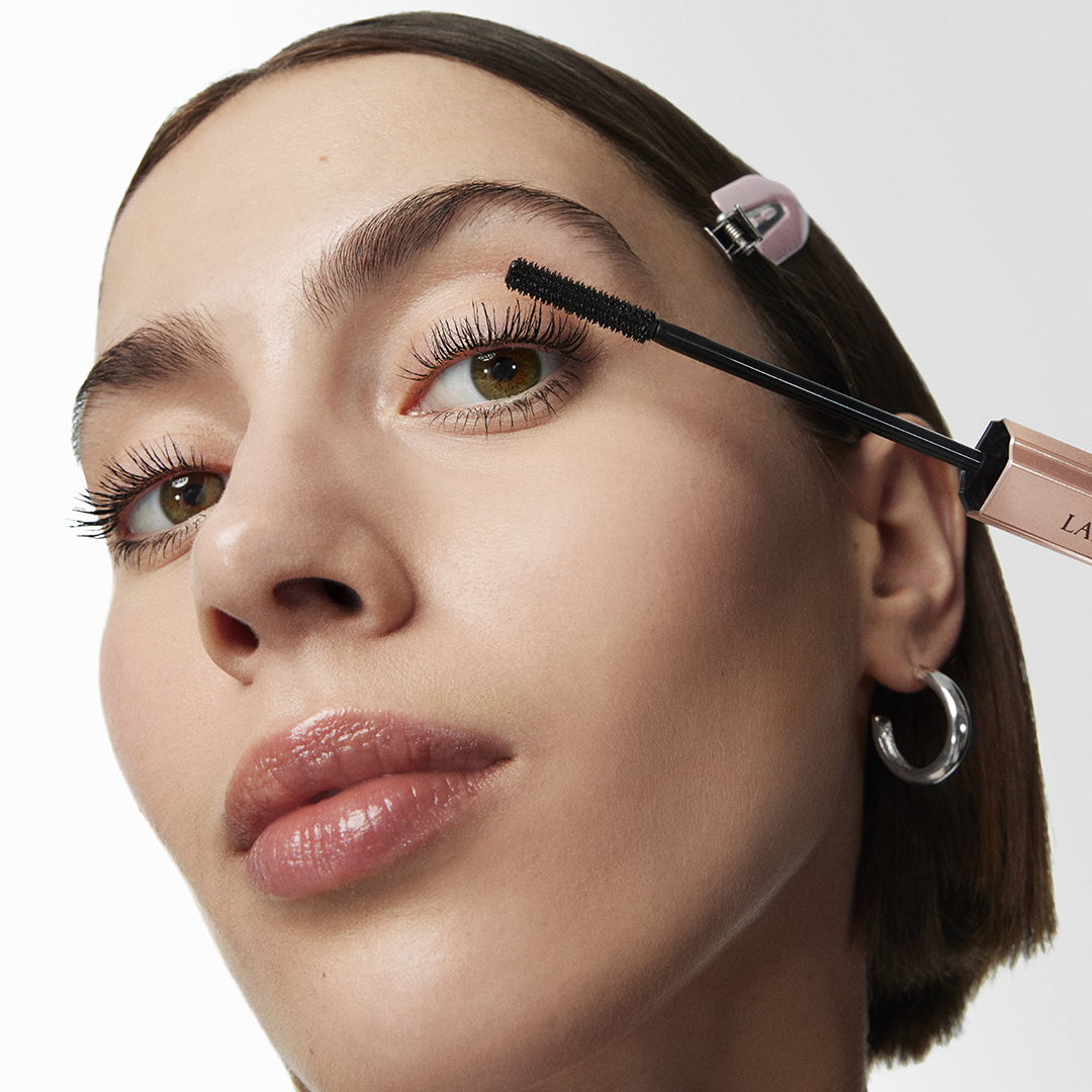 Lähikuva henkilöstä levittämässä Lancôme-ripsiväriä yläripsiin, selkeä harja ja avattu hylsy lähellä silmää.