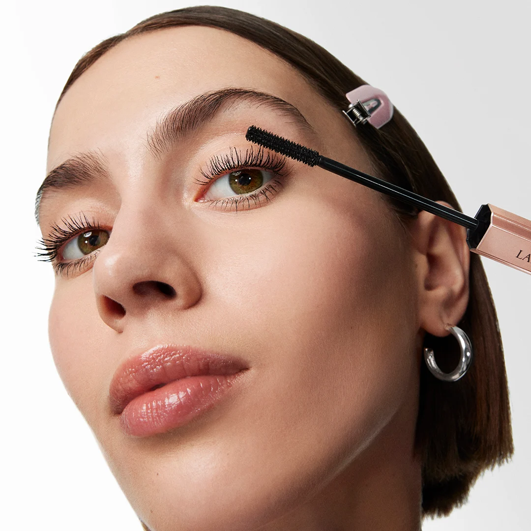 Lähikuva henkilöstä levittämässä Lancôme-ripsiväriä yläripsiin, selkeä harja ja avattu hylsy lähellä silmää.