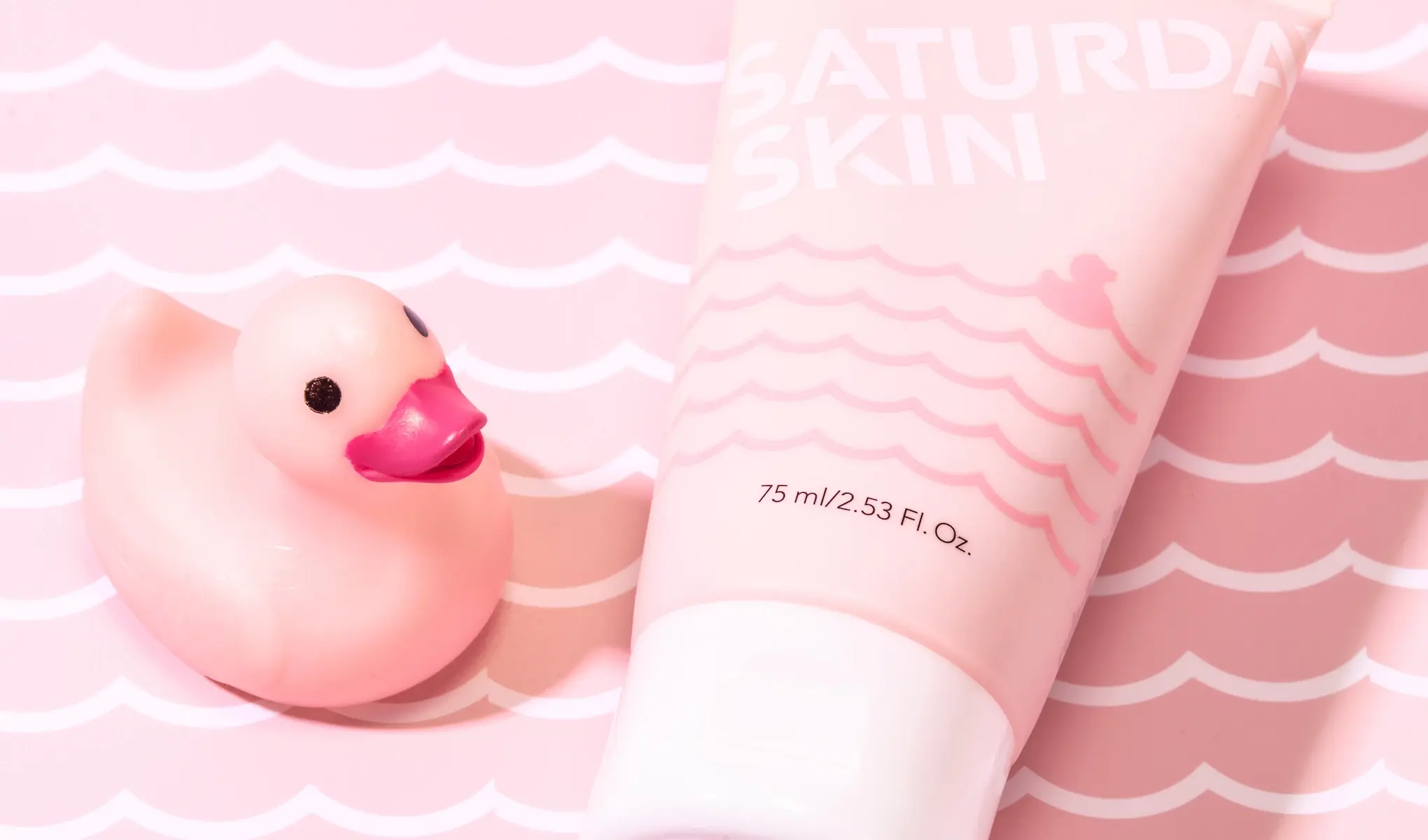 En rosa tube med hvit kork som inneholder hudpleieproduktet «Saturday Skin», er plassert på en rosa bakgrunn med hvite bølgemønstre. Ved siden av tuben står en liten rosa gummiand med rødt nebb og svarte øyne.