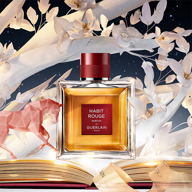 En flaska av Guerlain Habit Rouge Eau de Parfum mot en vit bakgrund. Flaskan är fyrkantig och genomskinlig, med en röd etikett som visar texten "Habit Rouge Eau De Parfum De Guerlain Paris". Korken är också röd och har en texturerad yta. Parfymens färg är gyllene, vilket ger en elegant och klassisk känsla.