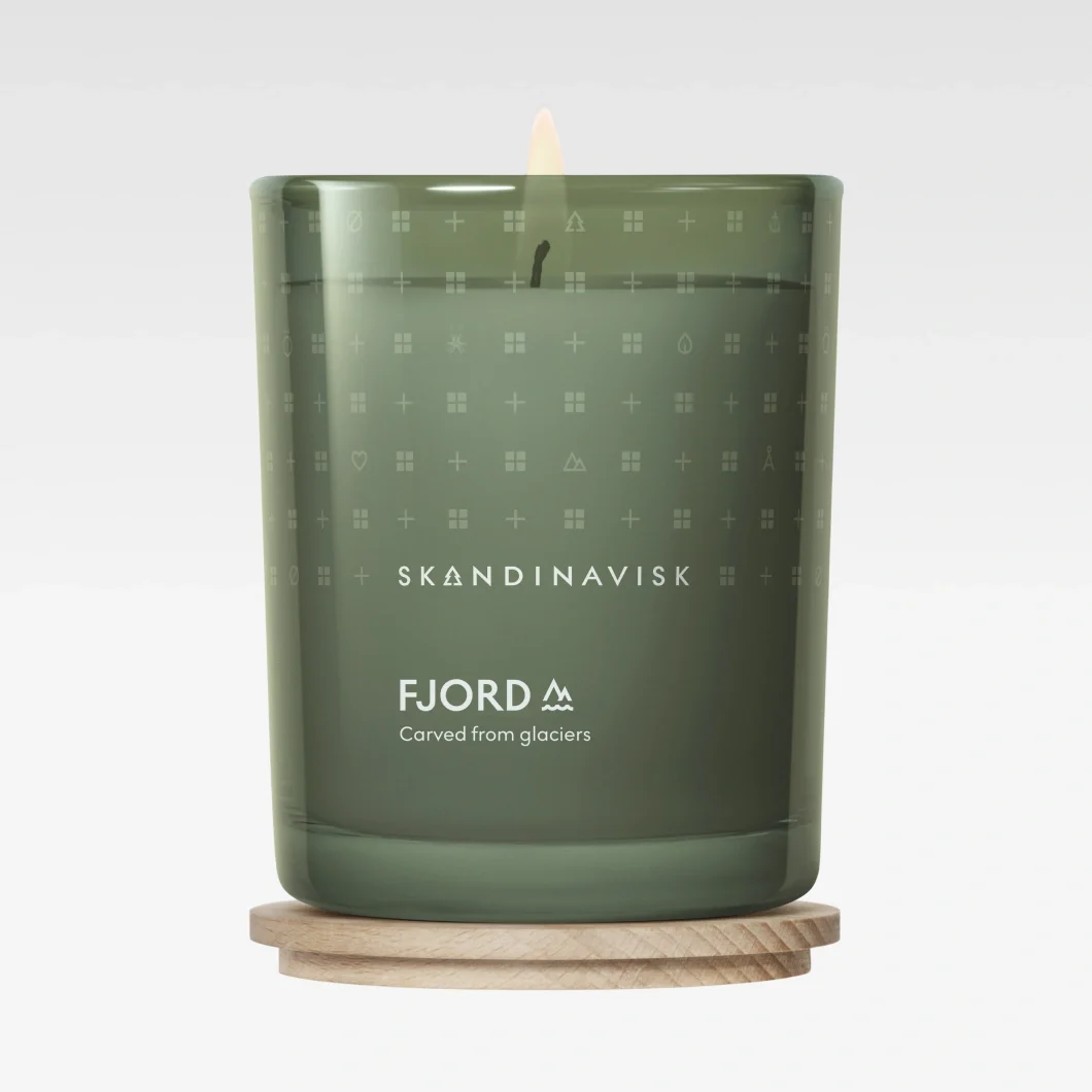 Grönt doftljus från SKANDINAVISK med texten “FJORD” och “Carved from glaciers” på glasbehållaren. Ljuset står på ett runt trälock mot ljusgrå bakgrund.