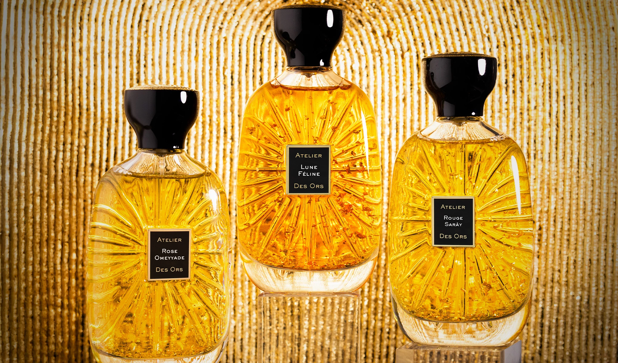 Tre parfymflaskor från Atelier Des Ors står uppradade mot en gyllene bakgrund. Flaskorna är fyllda med en guldfärgad vätska och har svarta korkar. Etiketterna på flaskorna visar namnen "Rose Omeyade", "Lune Féline" och "Rouge Sarây".