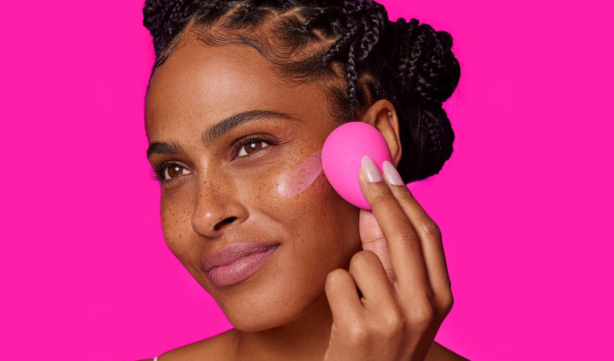 Person med flätat hår som håller en rosa sminksvamp mot sitt ansikte. Bakgrunden är starkt rosa, vilket matchar färgen på sminksvampen. Personen har välmanikyrerade naglar med ljusrosa nagellack.