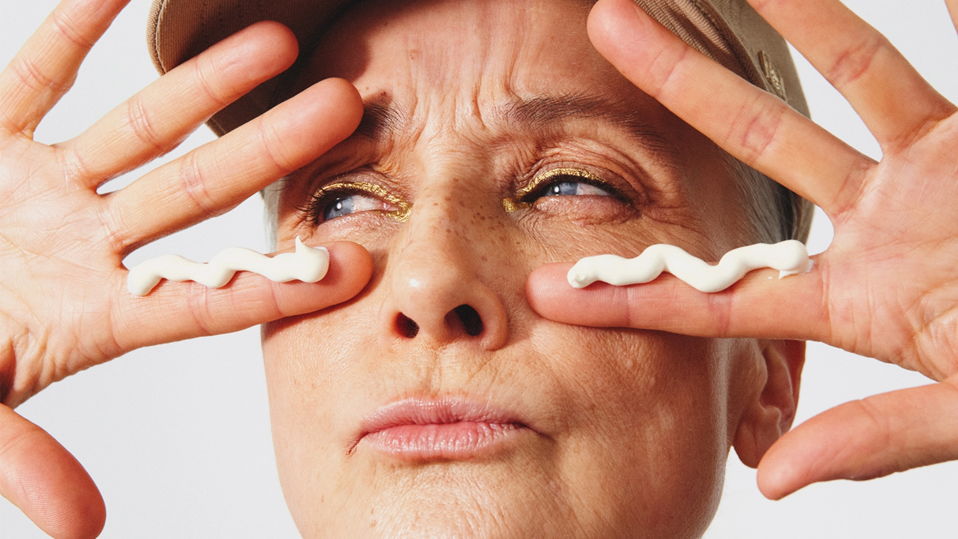 Porträtt av en person mot ljus bakgrund som håller båda händerna framför ansiktet. En ljus kräm är applicerad i vågform under båda ögonen. Personen bär keps och har smink med guldtonad ögonskugga. Fingrarna är utsträckta på vardera sidan av ansiktet.