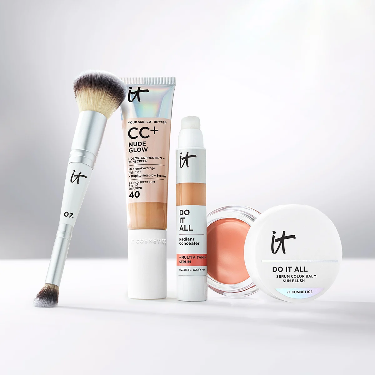 IT Cosmetics-sminkeprodukter mot lys bakgrunn, inkludert sminkekost, CC+ Nude Glow, Do It All Radiant Concealer og Do It All Serum Color Balm Sun Blush.